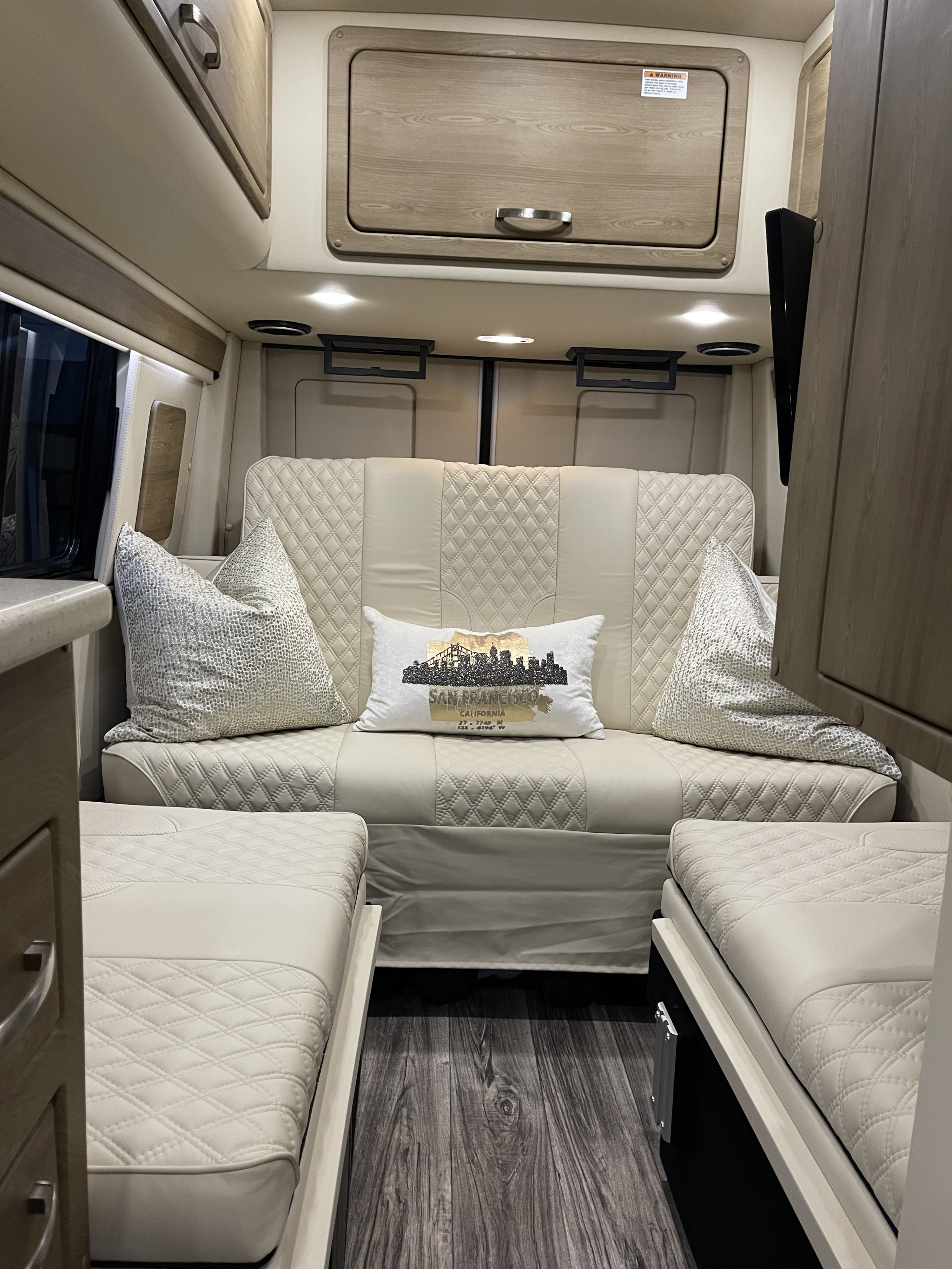 Modern & cozy RV rental interior: quilted sofa/beds, wood-look floor, and sleek overhead storage for your road trip comfort.. Midwest Automotive Designs Midwest Automotive Designs Class B 2022