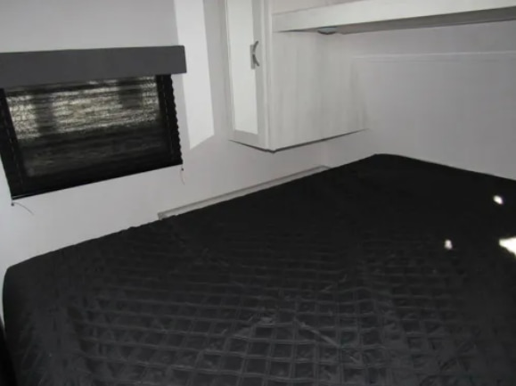 RV rental bedroom features a black quilted bed, window with blinds, and white overhead cabinets.. Forest River Tracer 2022