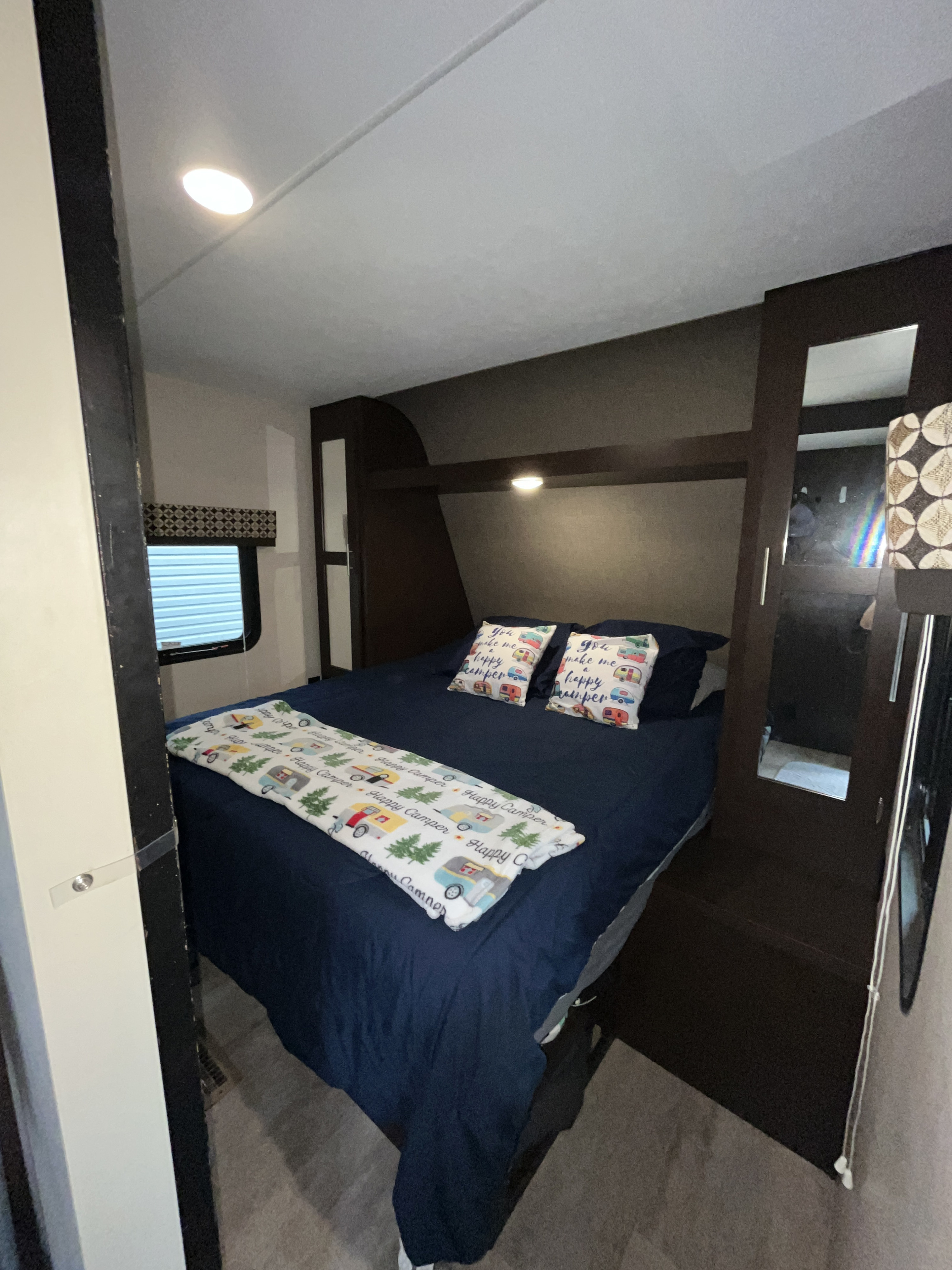 Inside a comfortable rv rental: a compact bedroom with a blue bed, "Happy Camper" themed pillows/blanket, and dark wood storage.. Sportsmen LE 2019