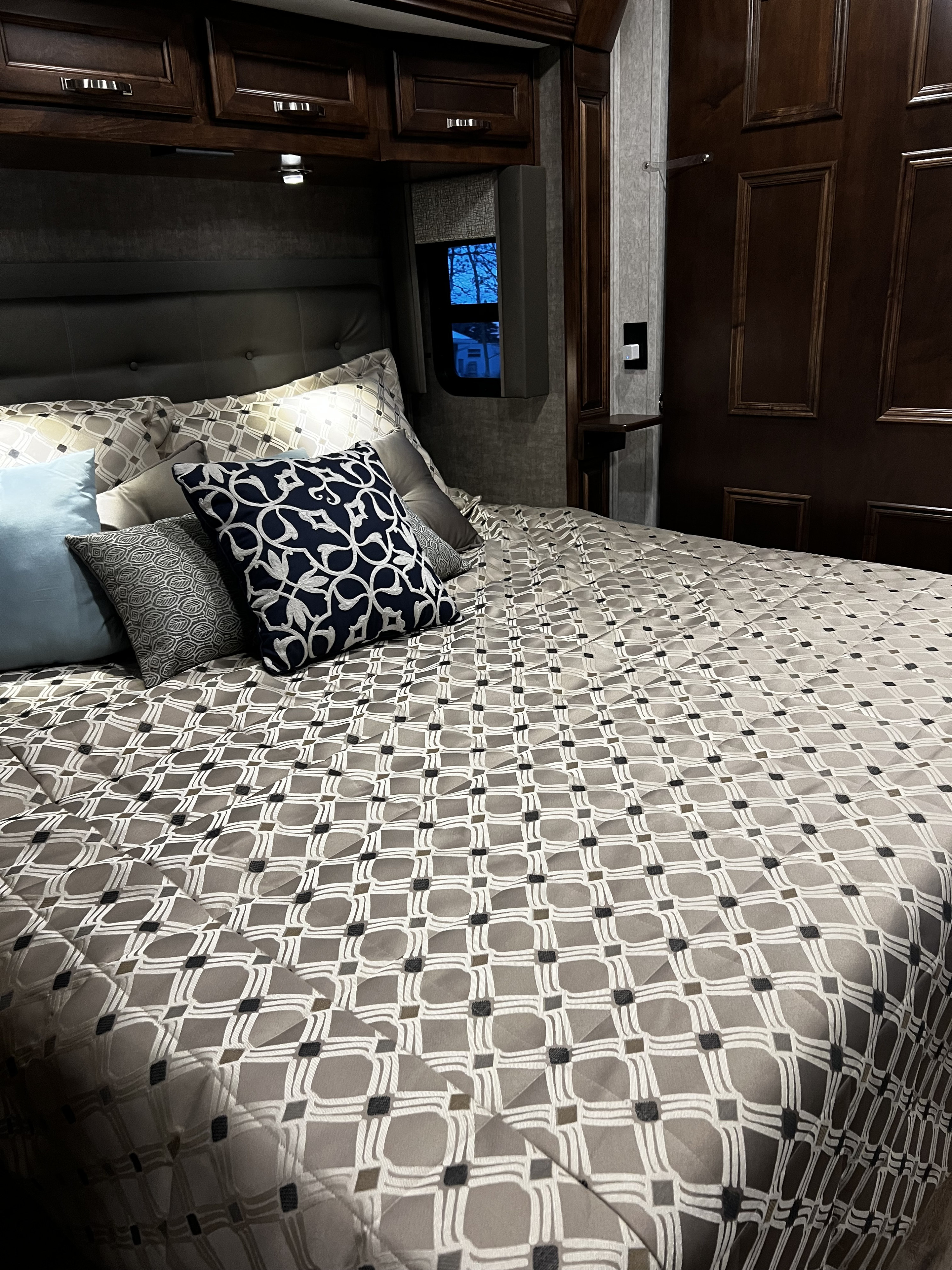 Cozy RV rental bedroom featuring a padded headboard, patterned comforter, decorative pillows, and wood cabinetry.. Renegade Custom Coaches Verona 2022