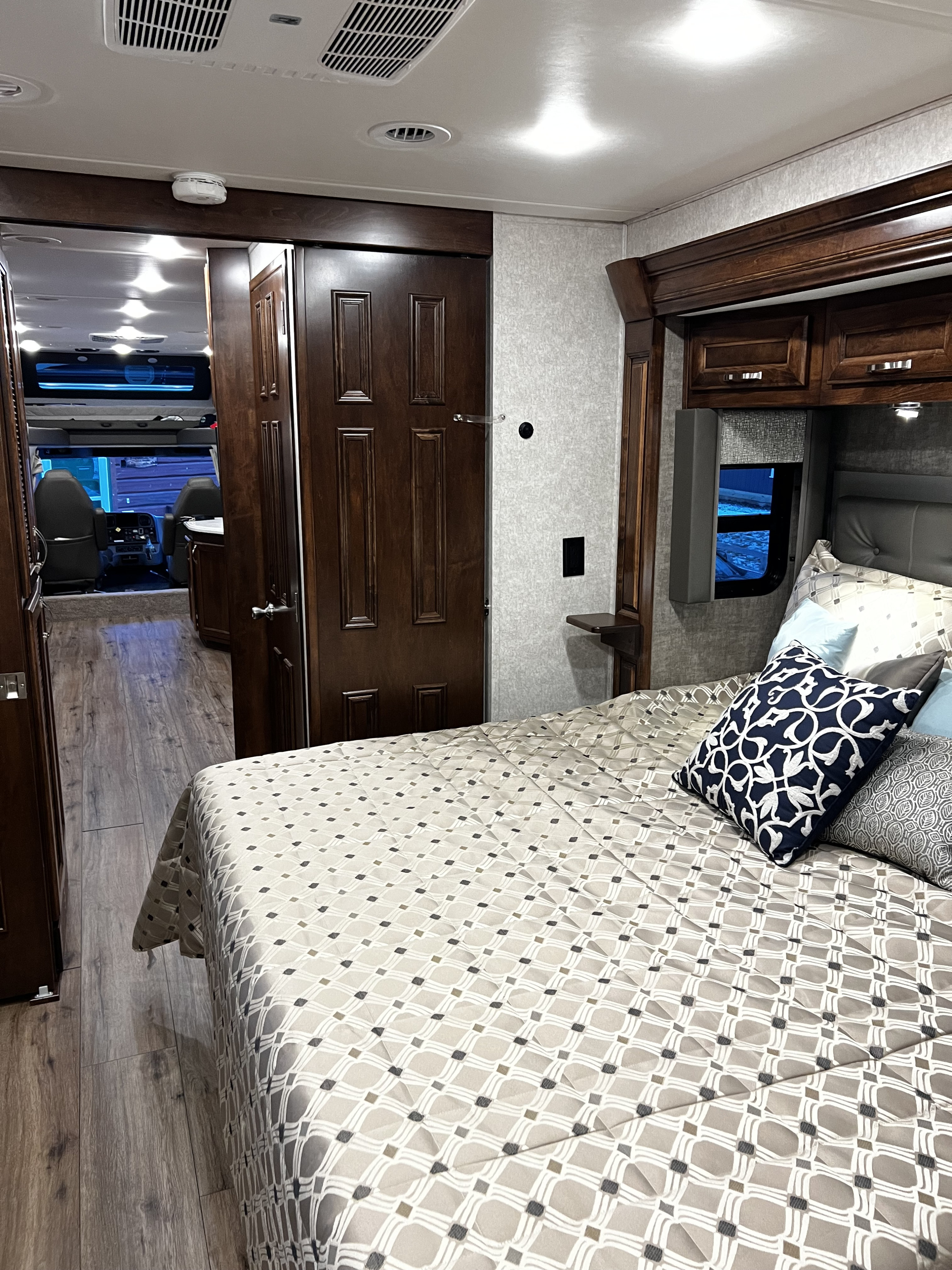 Cozy RV rental bedroom featuring patterned bedding, wooden cabinets, and a peek into the cockpit area for comfortable travel.. Renegade Custom Coaches Verona 2022