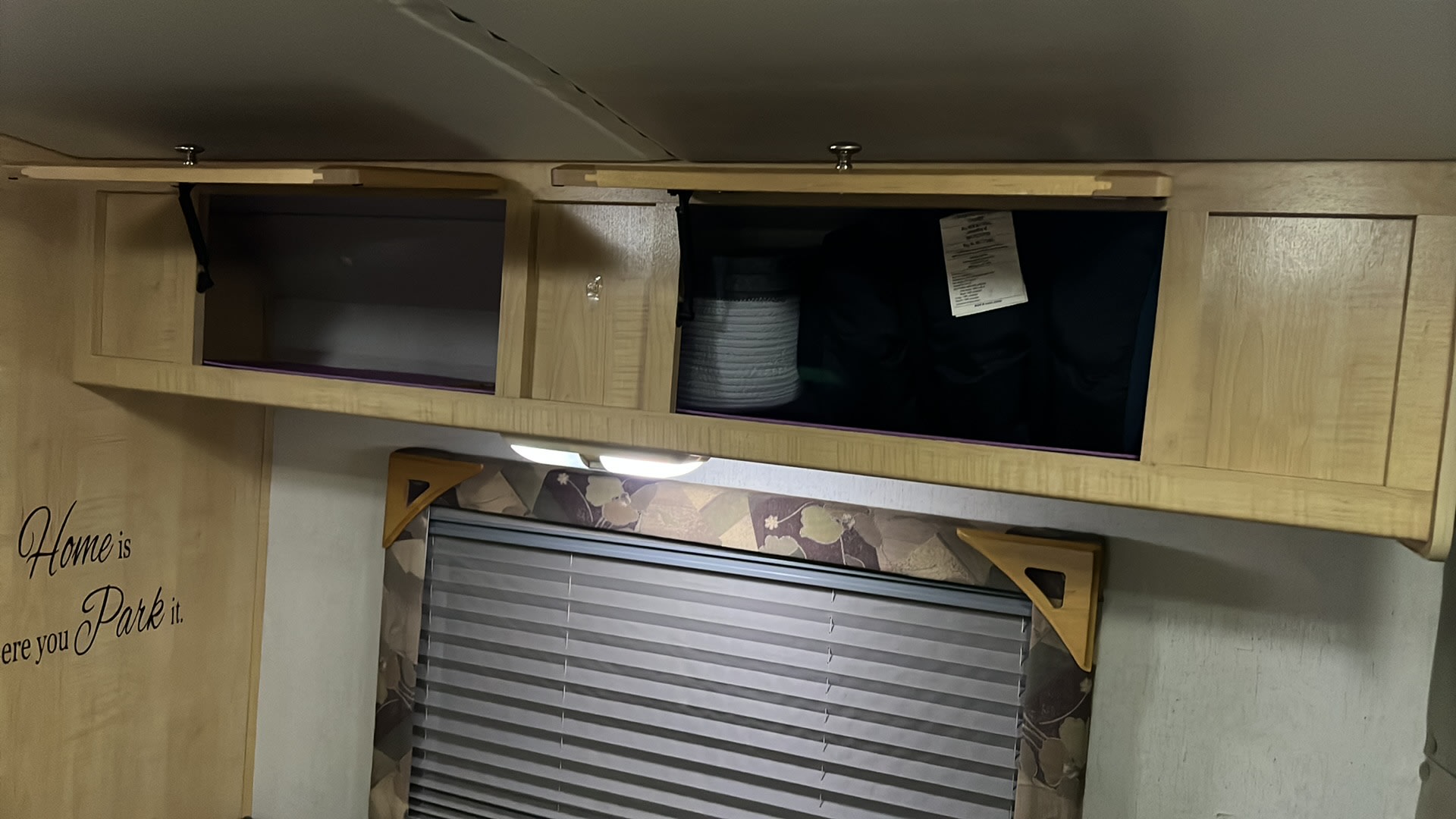 Cozy RV rental interior with wooden overhead storage cabinets, one open, a window with blinds, and a "Home is where you Park it" decal.. R-Vision Trail-Lite B-Plus 2004