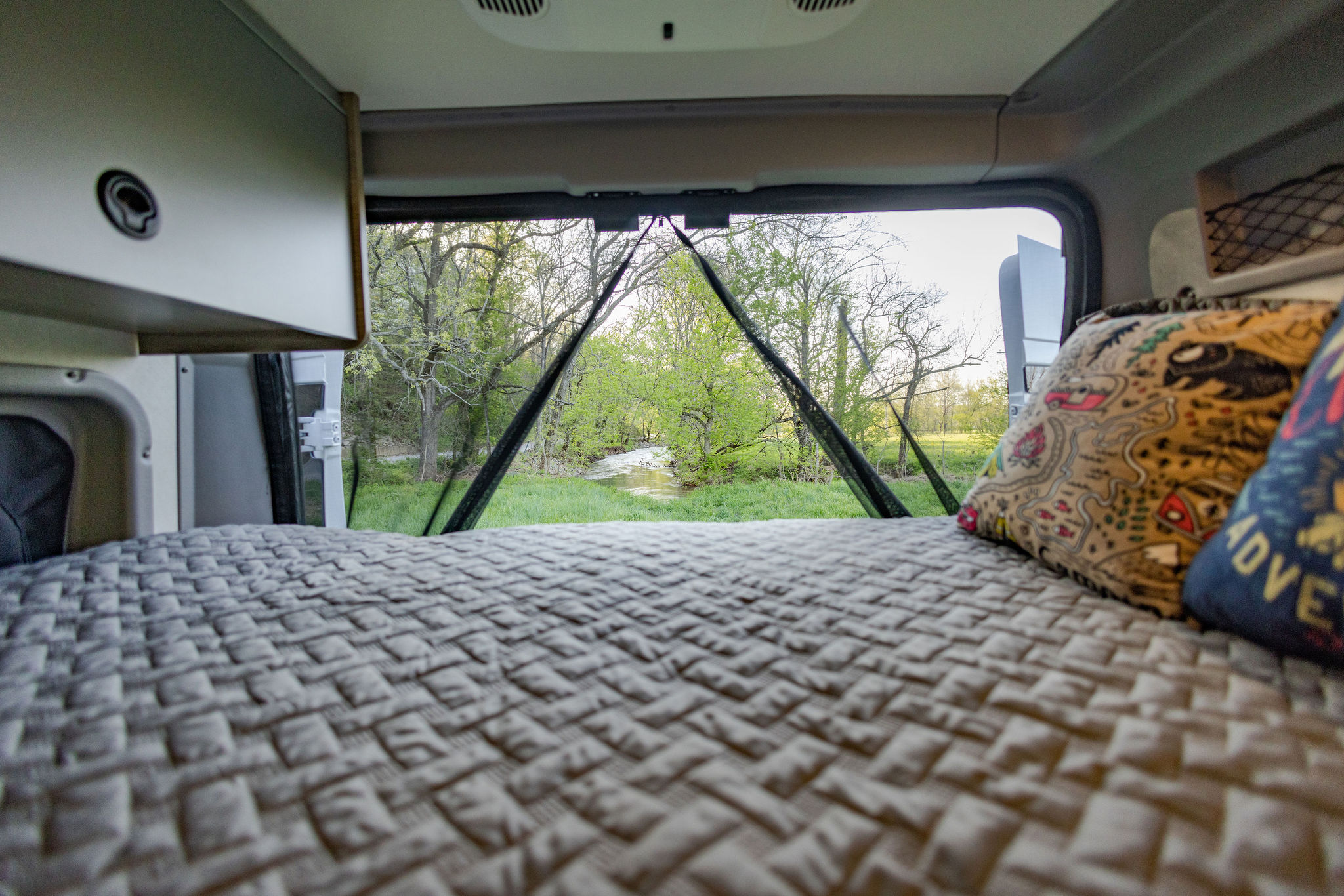 RV rental: Comfy bed, pillows, and open back doors reveal a serene view of a stream flowing through lush green trees.. Winnebago Solis Pocket 2022