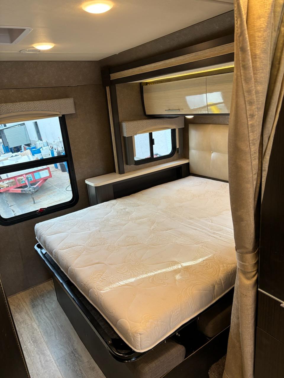 Cozy RV rental bedroom showcasing a clean mattress, soft ambient lighting, overhead storage, and large windows.. Thor Vegas 2018