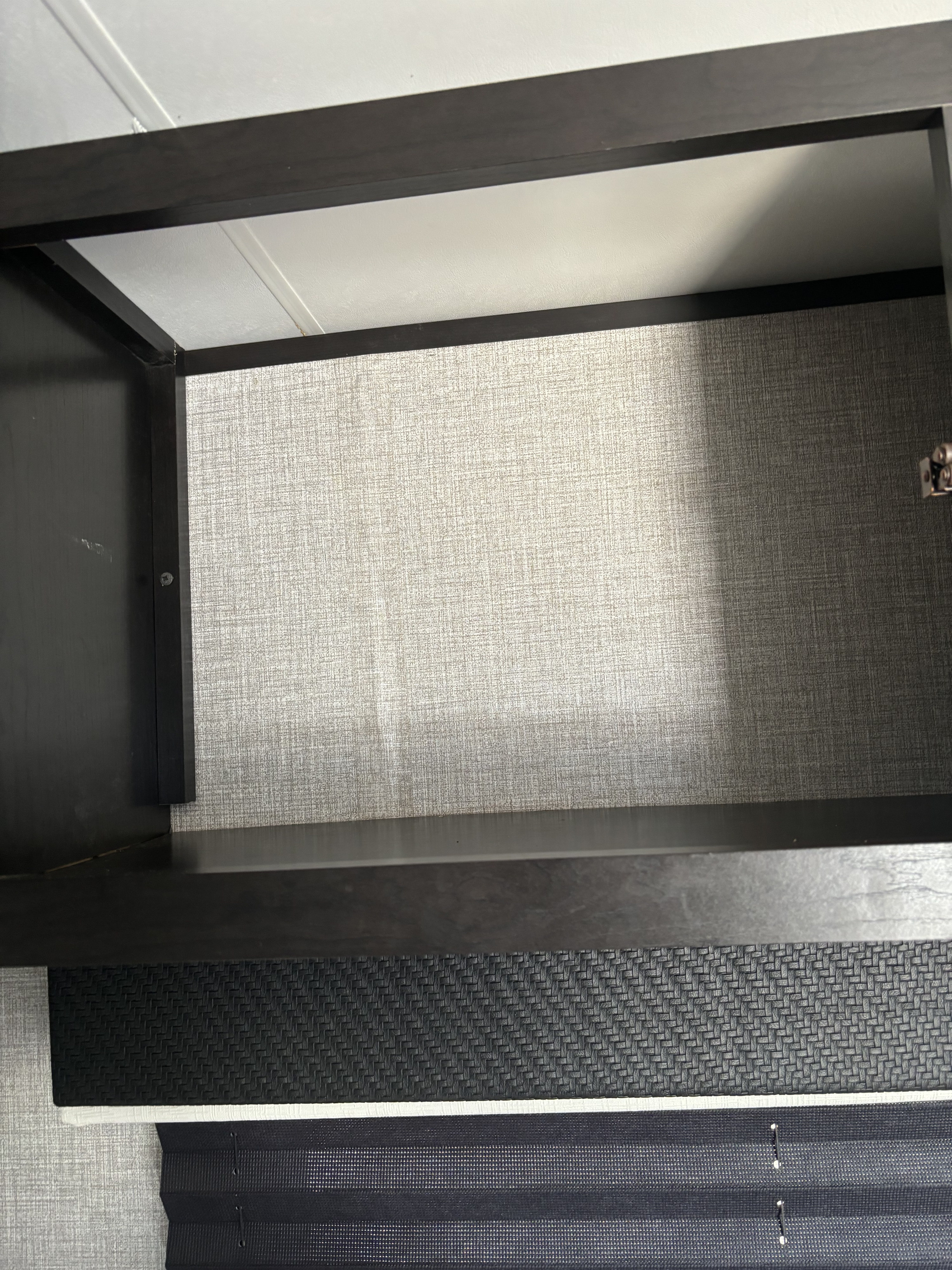Inside an RV rental, showing an empty cabinet with a textured back wall and dark shelves, above a pleated window blind.. Jayco Jay Flight SLX Baja Edition 2022