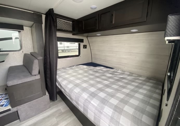 RV rental interior features a bed with plaid bedding, adjacent dinette seating, and dark overhead storage.. Jayco Jay Flight SLX Baja Edition 2023