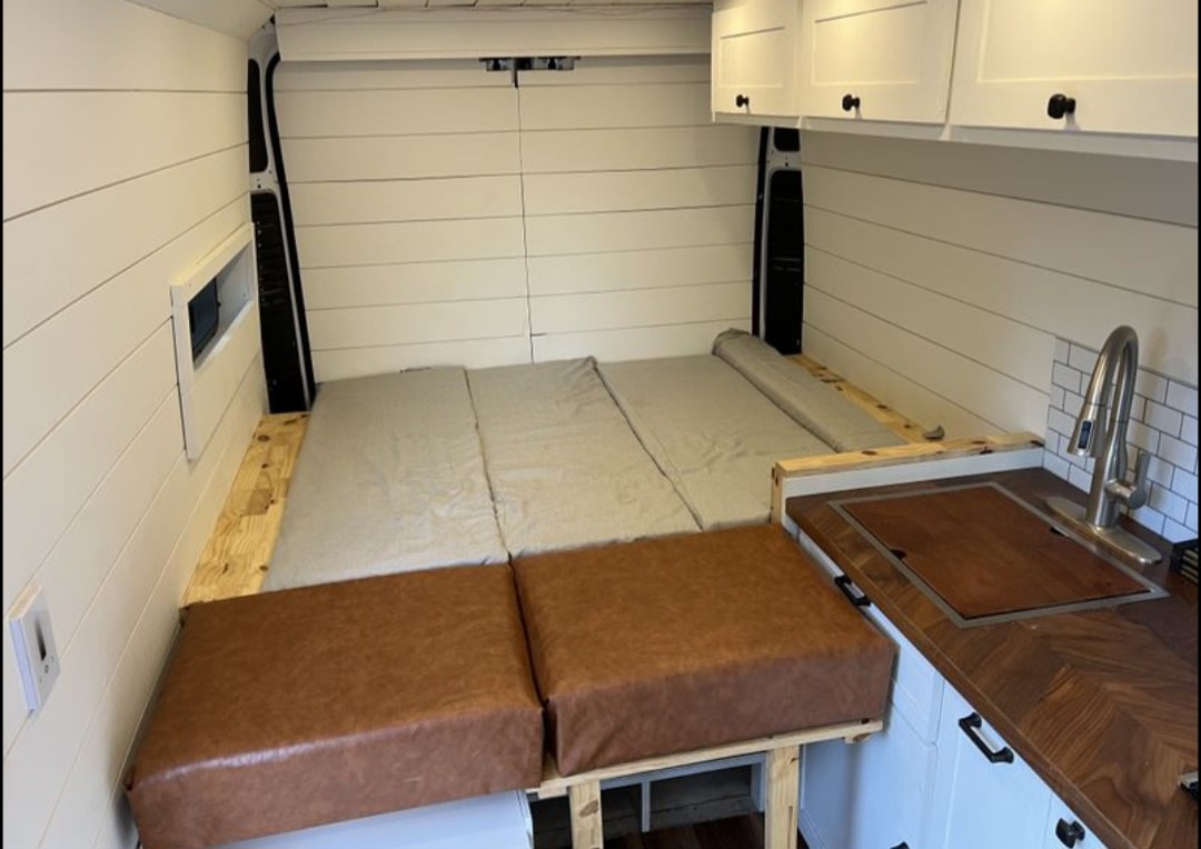 Inside an RV rental camper van: white shiplap walls, bed, seating, and a compact kitchen with a sink. Ready for road trips!. Ram Promaster 2019