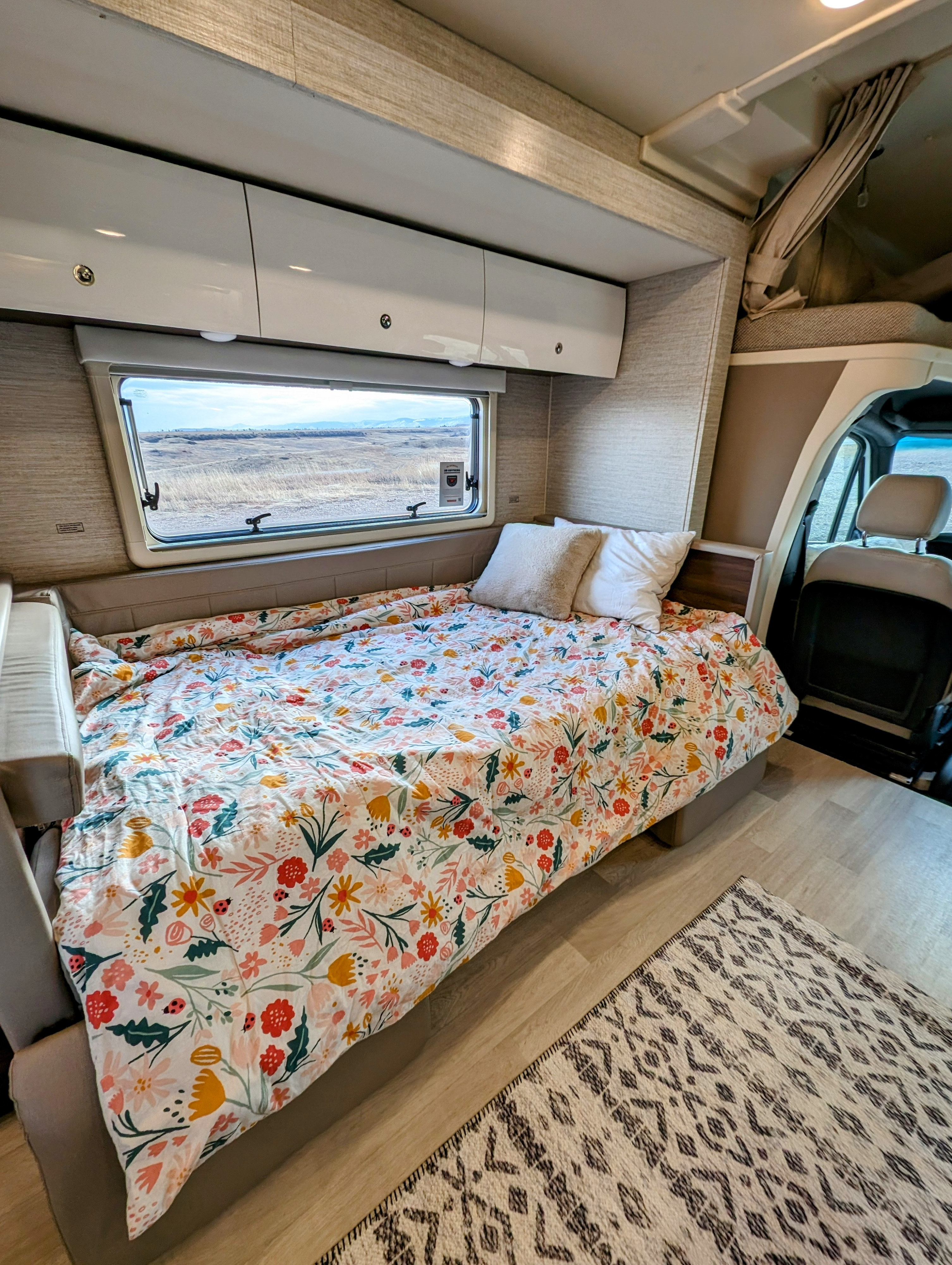 Your comfy RV rental awaits! Cozy bed, floral duvet, scenic window view of arid land, overhead storage, and patterned rug.. Winnebago Navion 2023