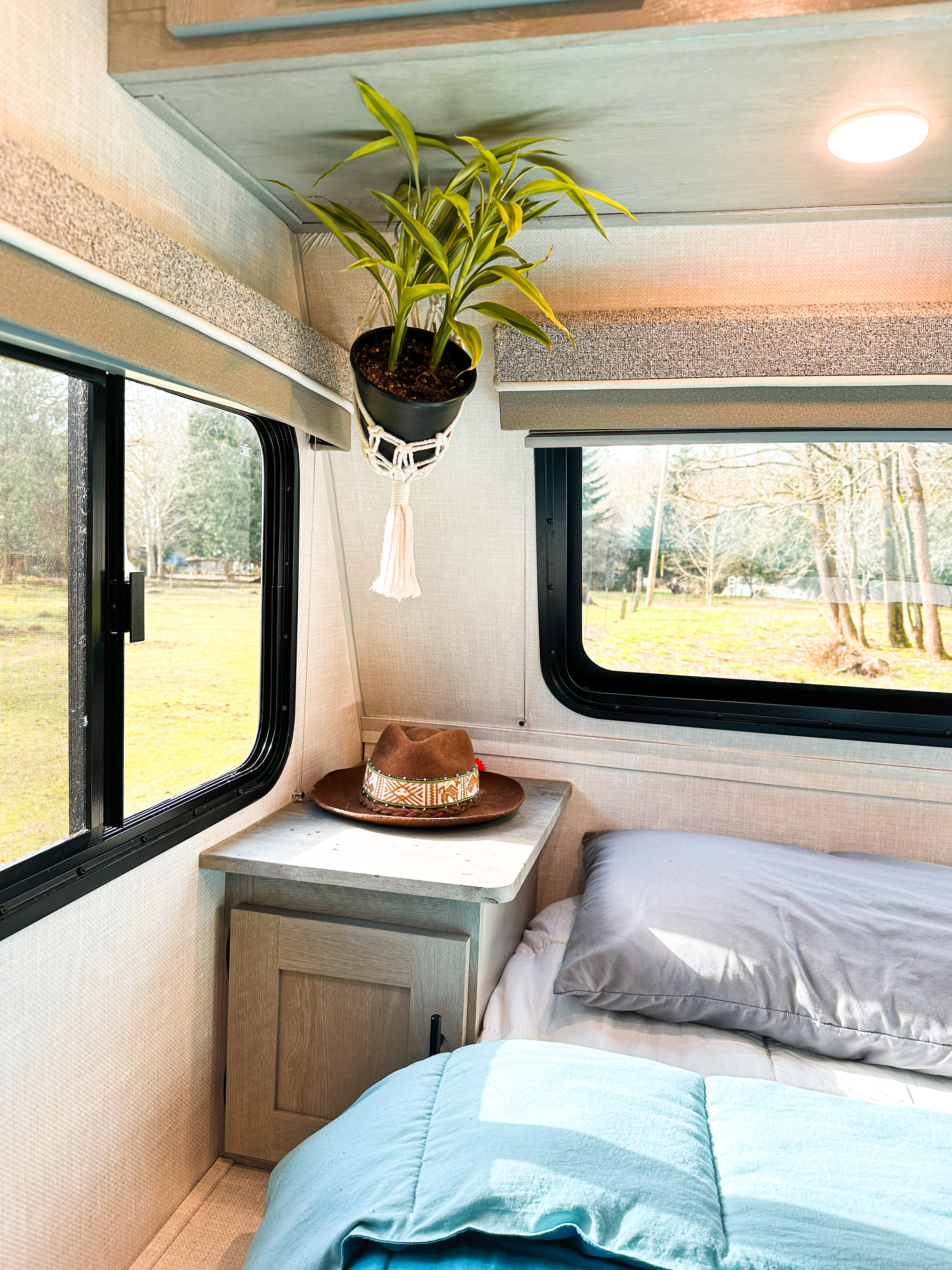 Cozy RV rental bedroom with bed, blue/grey bedding, nightstand with hat, hanging plant & windows to nature.. Forest River Ibex 2021