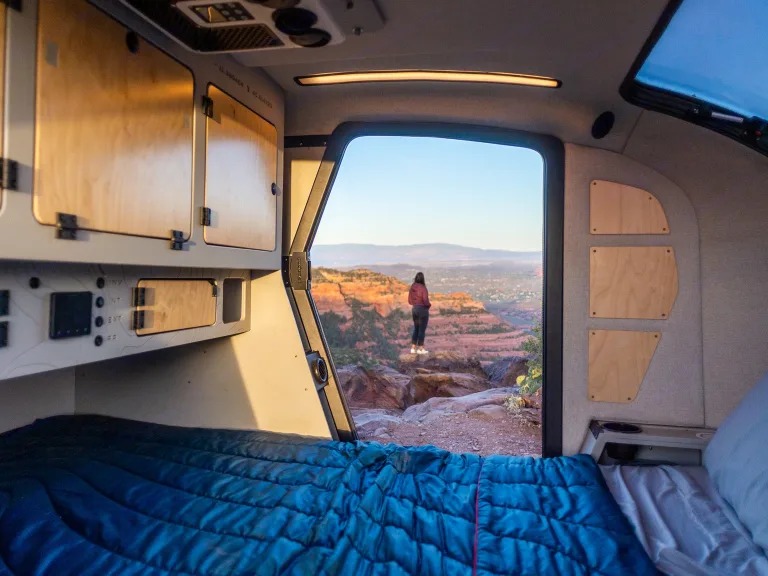 An RV rental provides a cozy bed with epic views! A person looks out at red rock canyons from inside a camper.. Other Other 2024