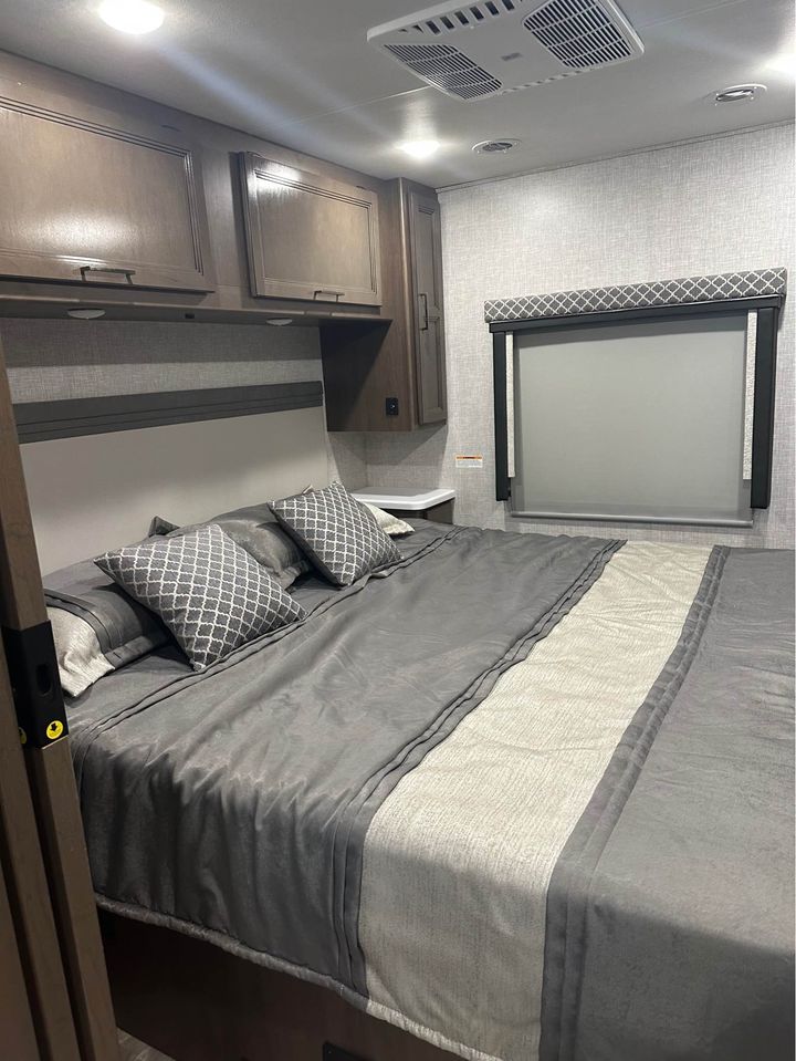 RV rental bedroom: a comfortable gray bed, wood cabinets, and window with patterned valance under a bright ceiling. A cozy interior.. Thor Windsport 2023