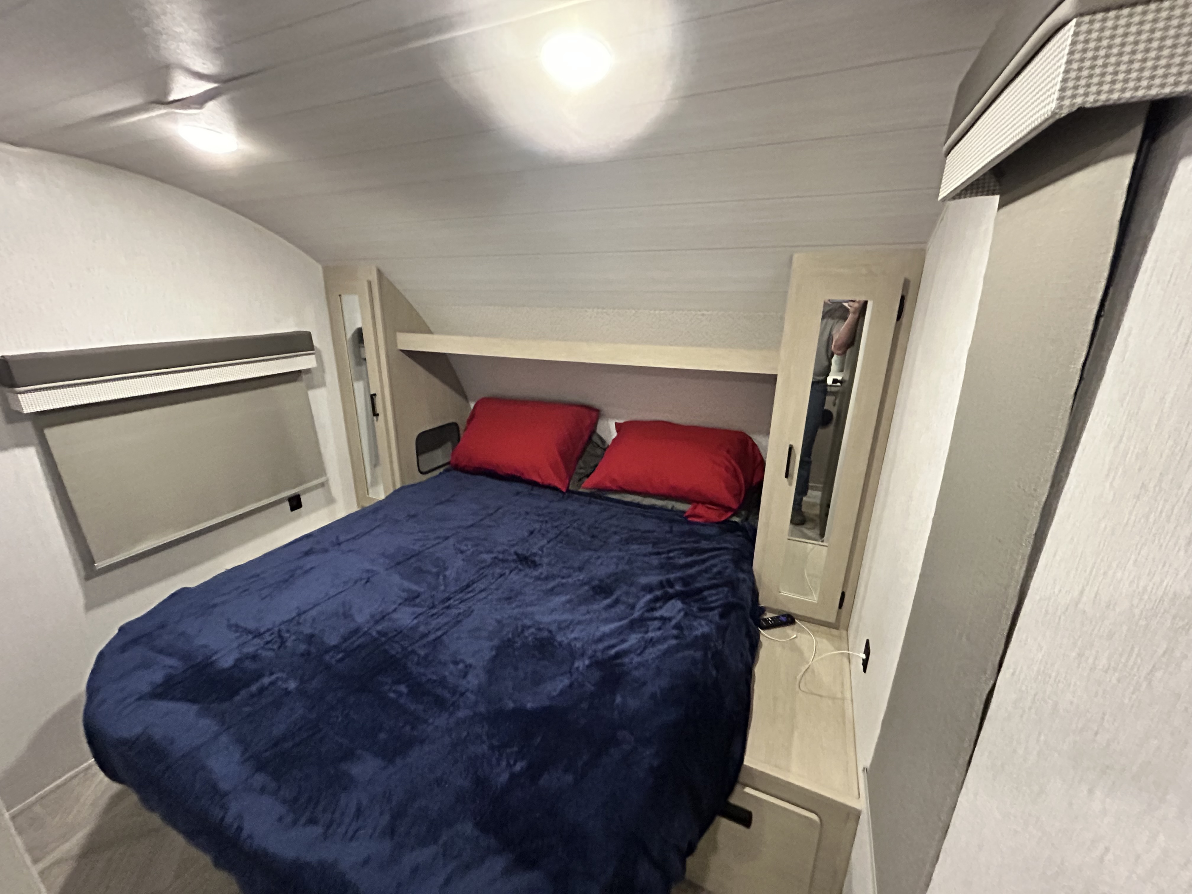 This compact RV rental bedroom features a blue comforter, red pillows, overhead shelf, and mirrored wardrobes for storage.. Forest River Wildwood X-Lite Platinum 2023