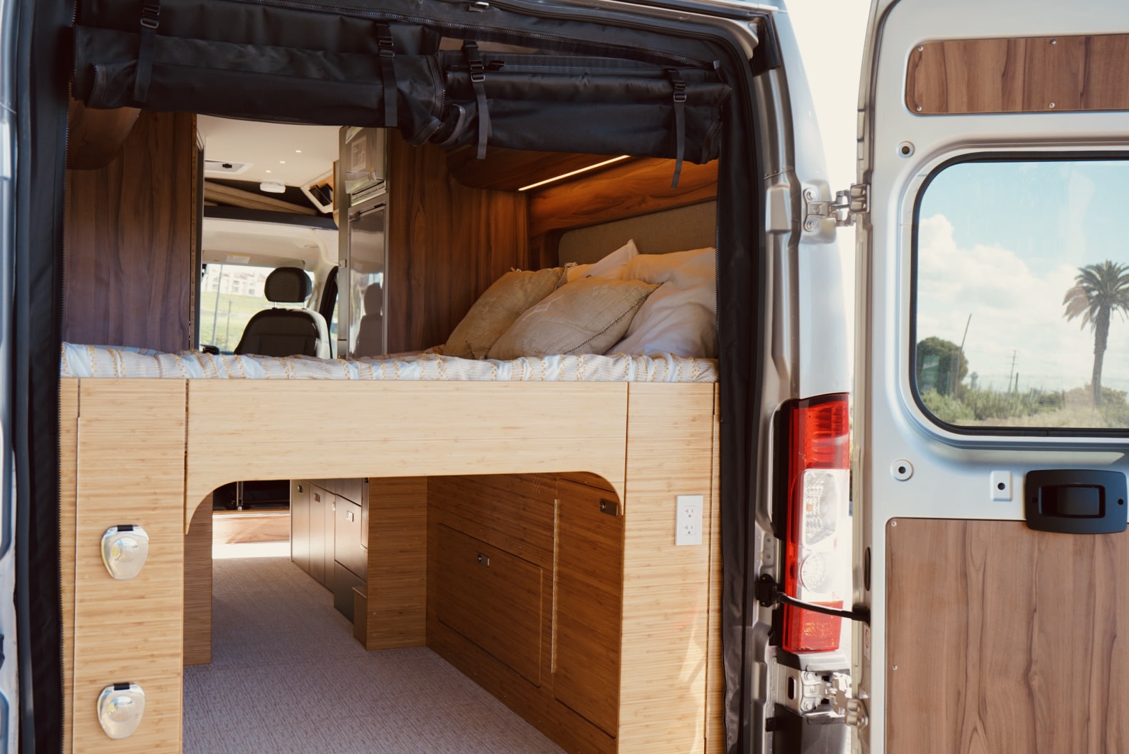 View from the back of an RV rental: a raised bed, modern kitchen, and front seats inside. Open doors reveal a palm tree outside.. Noovo Lite 2023