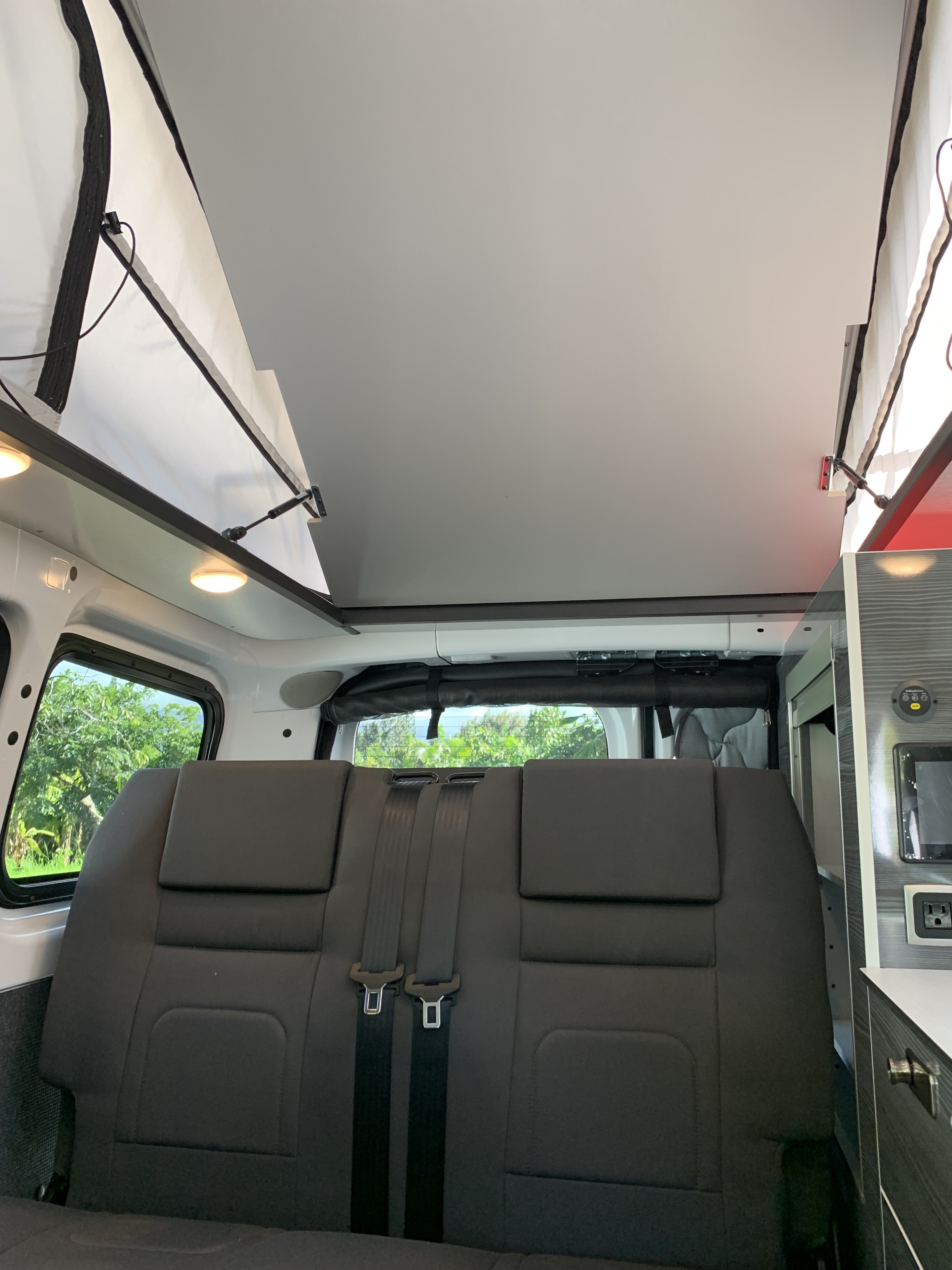 **RV rental interior**: Shows a raised pop-top roof, dark grey bench seats with seatbelts, and windows looking out to lush trees.. NIssan Envy 2021