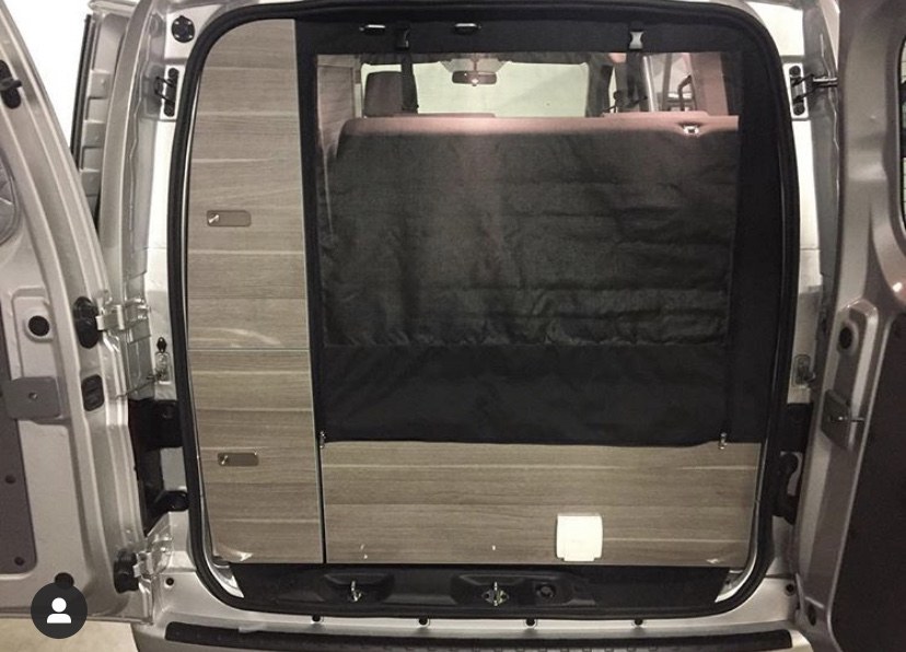 View from rear of RV rental showing wood-paneled interior, dark bed/storage area, and functional camper van setup.. NIssan Envy 2021