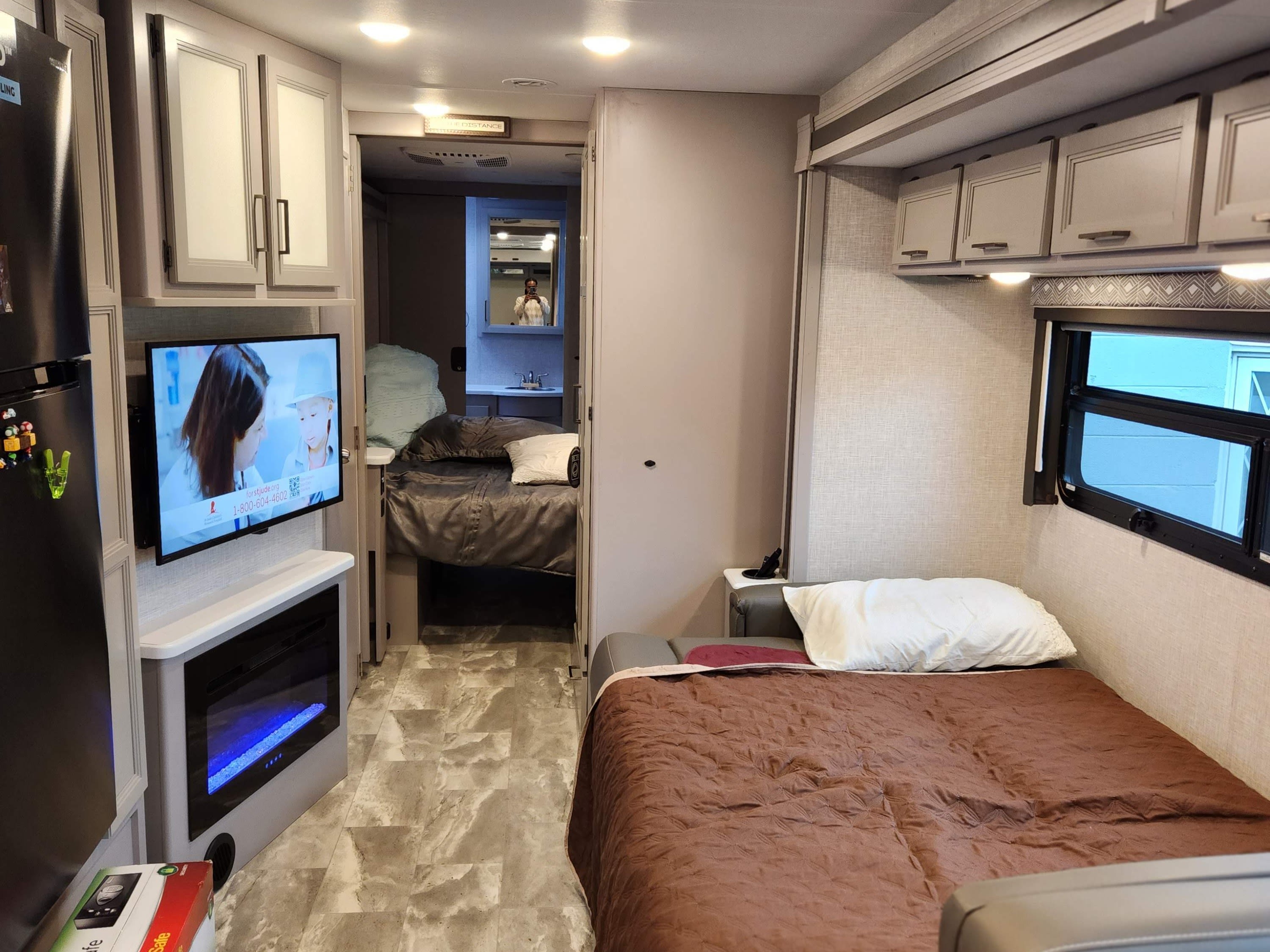 Modern RV rental interior featuring a main bed, TV with fireplace, refrigerator, and a glimpse of the rear bedroom/bathroom.. Thor Windsport 2023