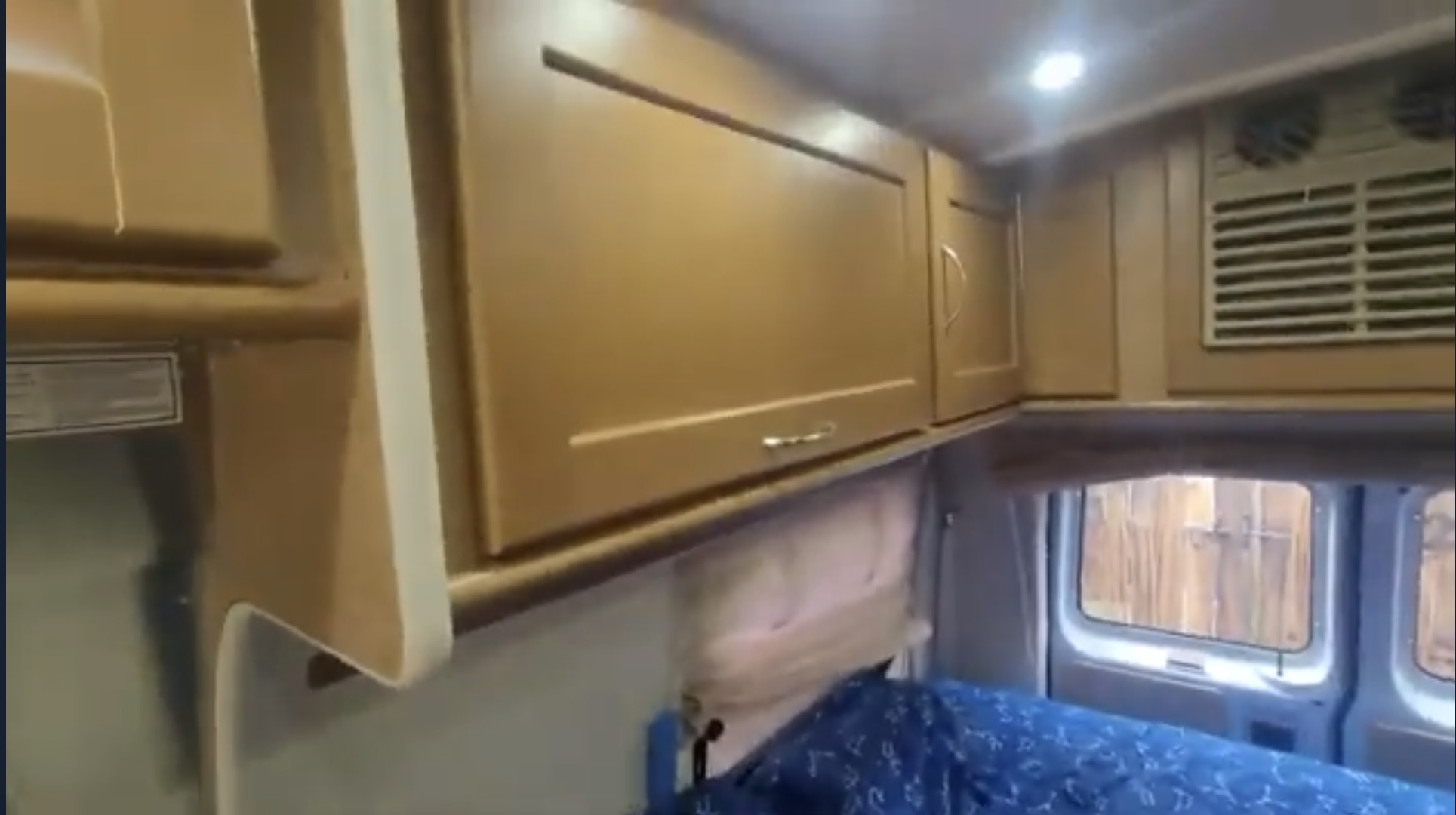 Inside an RV rental: light wood overhead cabinets, bright ceiling lights, a blue patterned bed below, and rear windows showing a fence.. Pleasure-Way Excel TS 2012