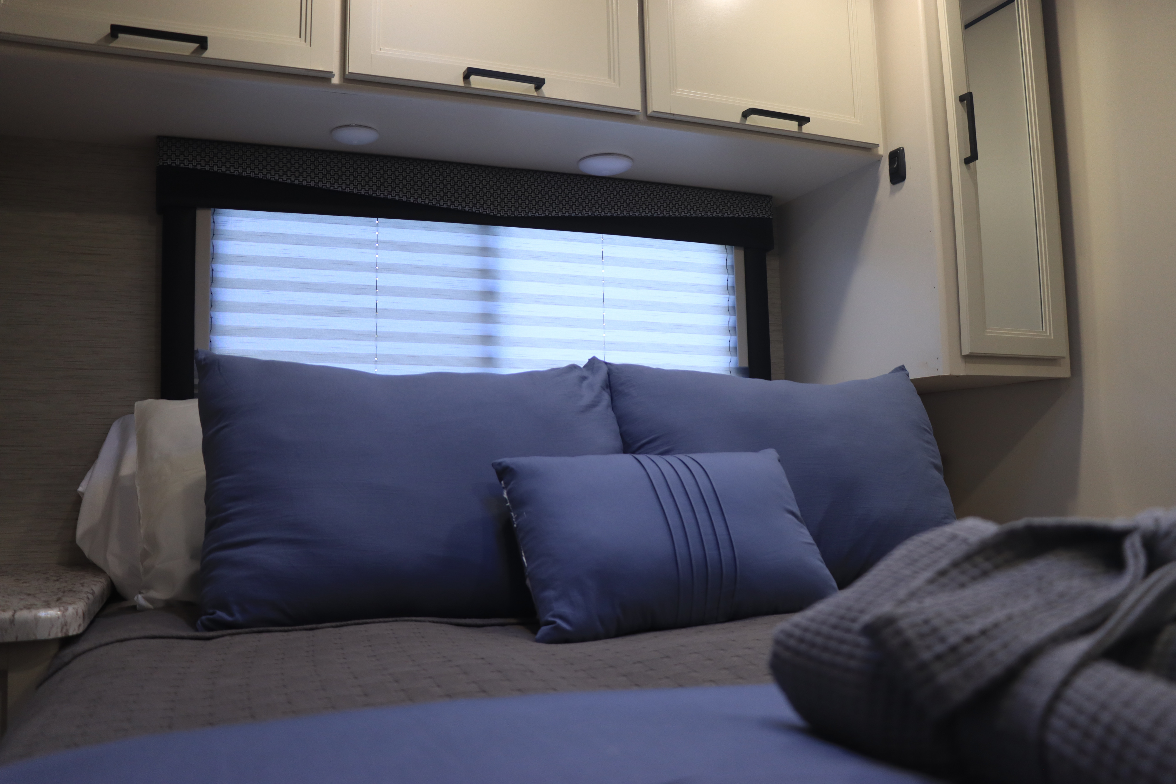 Serene RV rental bedroom featuring a cozy bed, blue pillows, overhead cabinets, and a bright window with blinds.. Thor Freedom Elite 2023