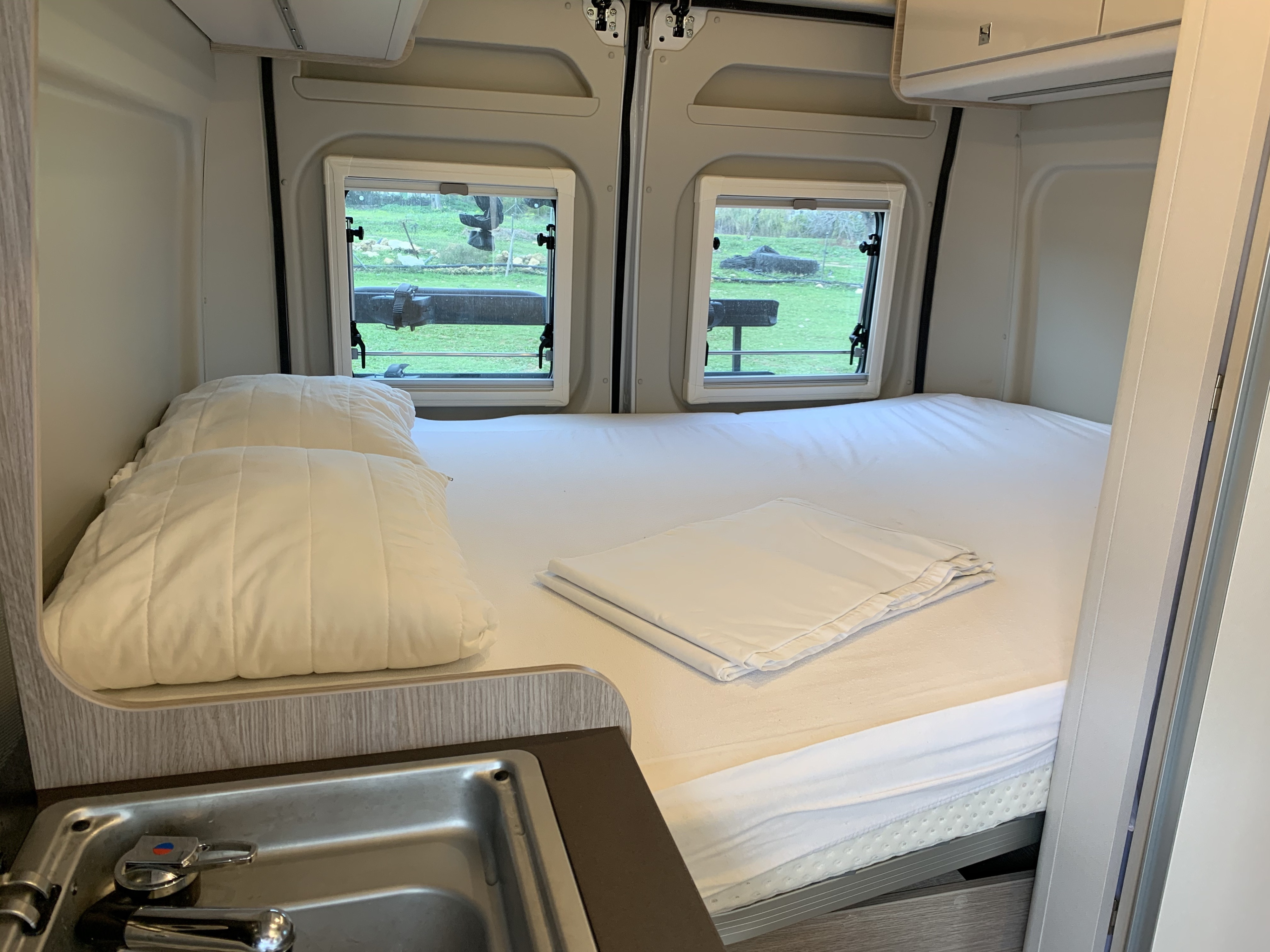 Inside this RV rental: a pristine bed with white linens, large windows showcasing greenery, and a practical sink area.. BURSTNER Campeo 2026