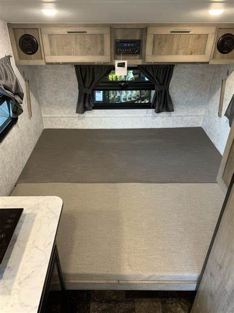 View inside an RV rental showing a light grey cushioned bed, overhead cabinets, a window, and control panel.. Prolite E-Volt 2021