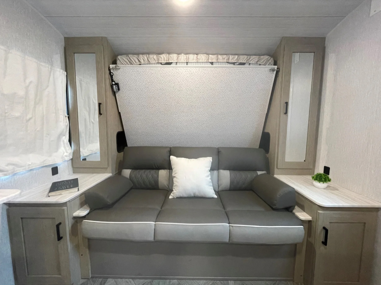 Cozy rv rental interior with a gray convertible sofa/bed and a stowed murphy bed, flanked by mirrored cabinets.. Forest River Wildwood X-Lite 2021