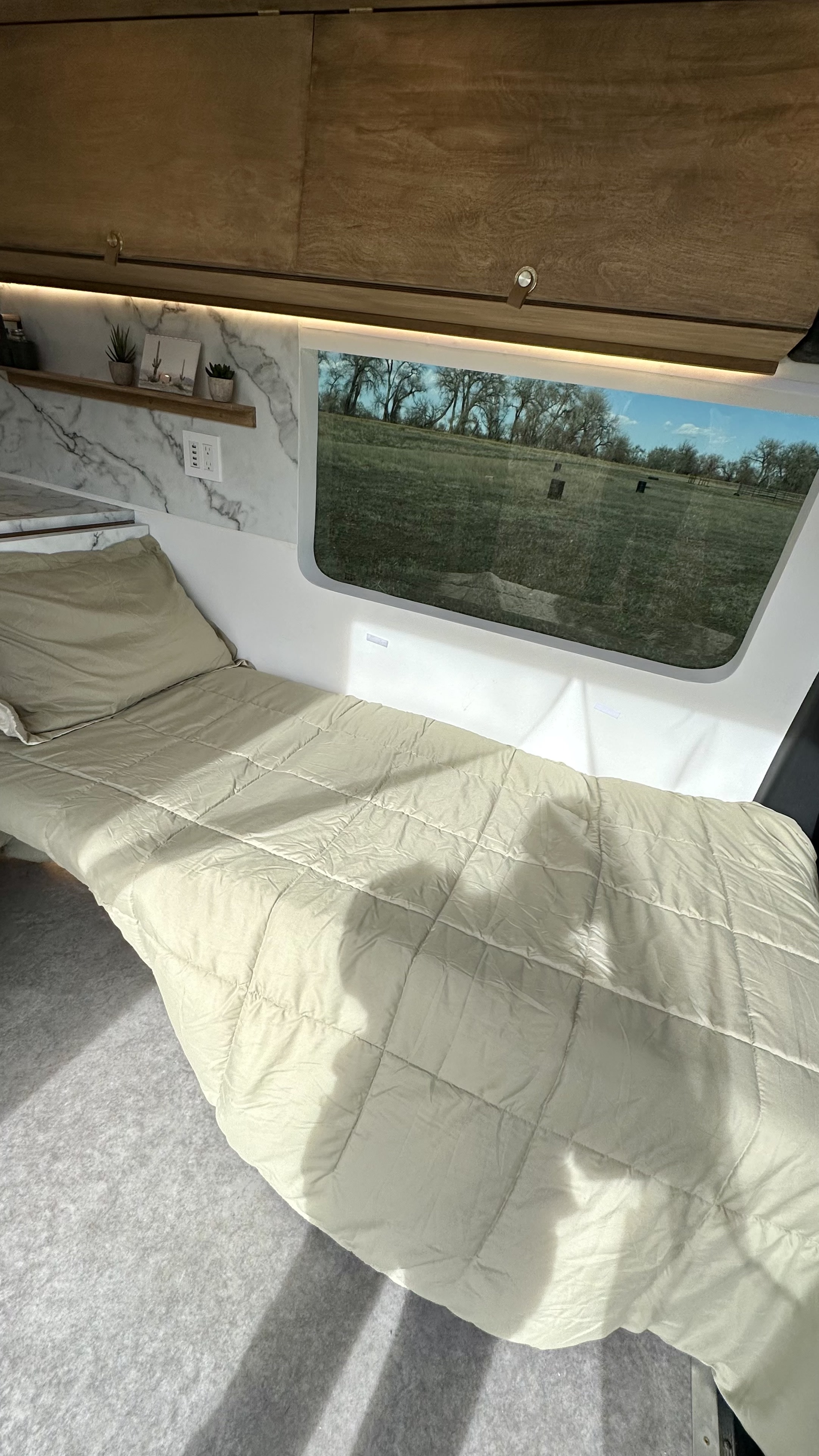RV rental sleeping area: a comfortable bed with a scenic window showing a grassy field and trees.. Mercedes-Benz Sprinter High Roof Medium wheelbase 2021