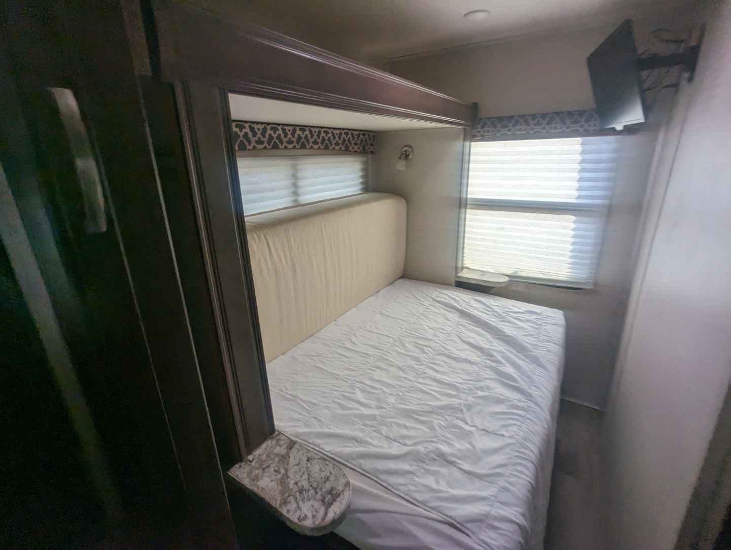 This RV rental bedroom has a cozy bed with padded headboard, window with blinds, and a wall-mounted TV.. Jayco Redhawk 2019
