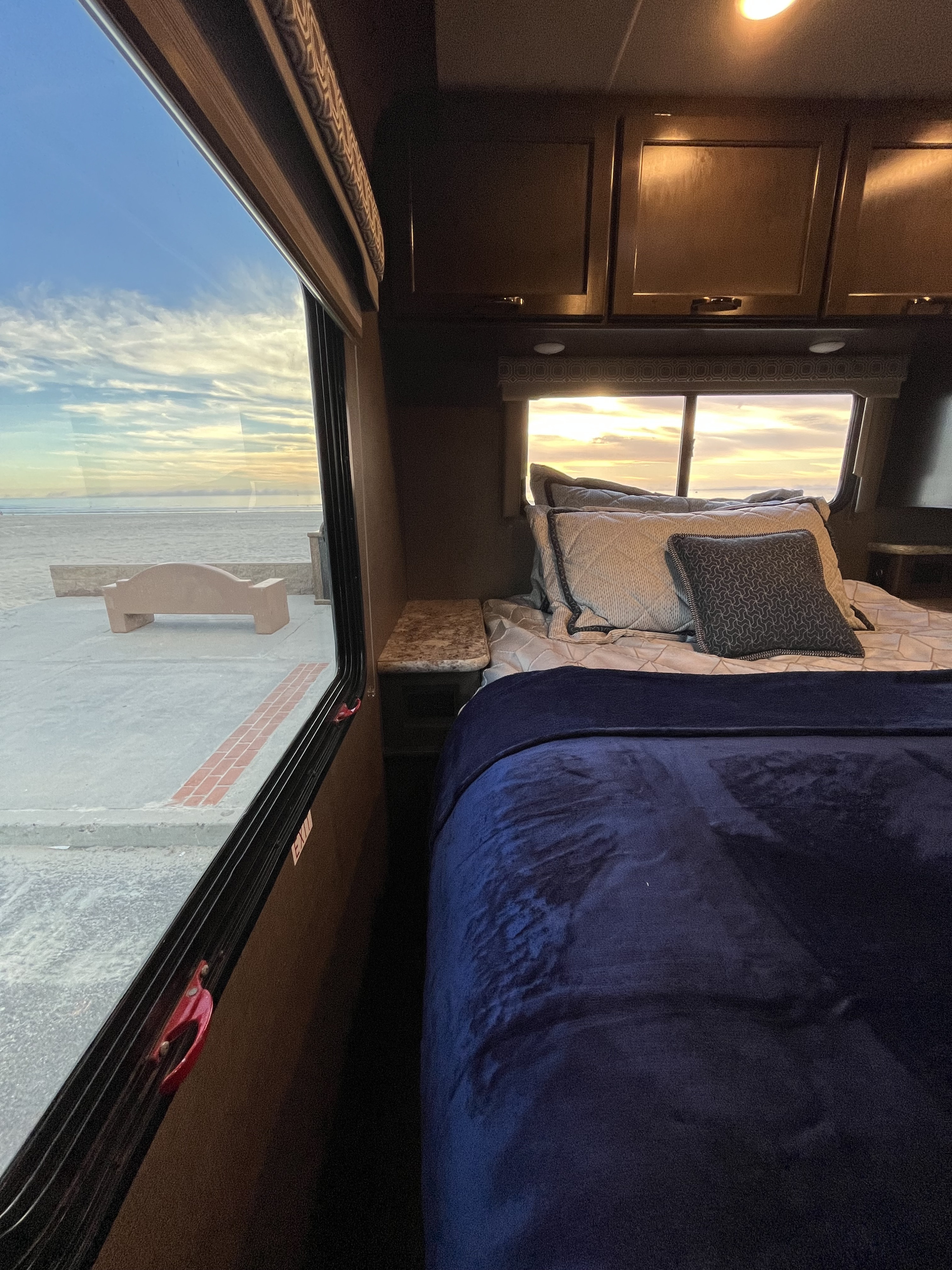 Inside an RV rental: A cozy bedroom with a blue blanket, pillows, and a window offering a beautiful ocean sunset view.. Thor Motor Coach Freedom Elite 2018