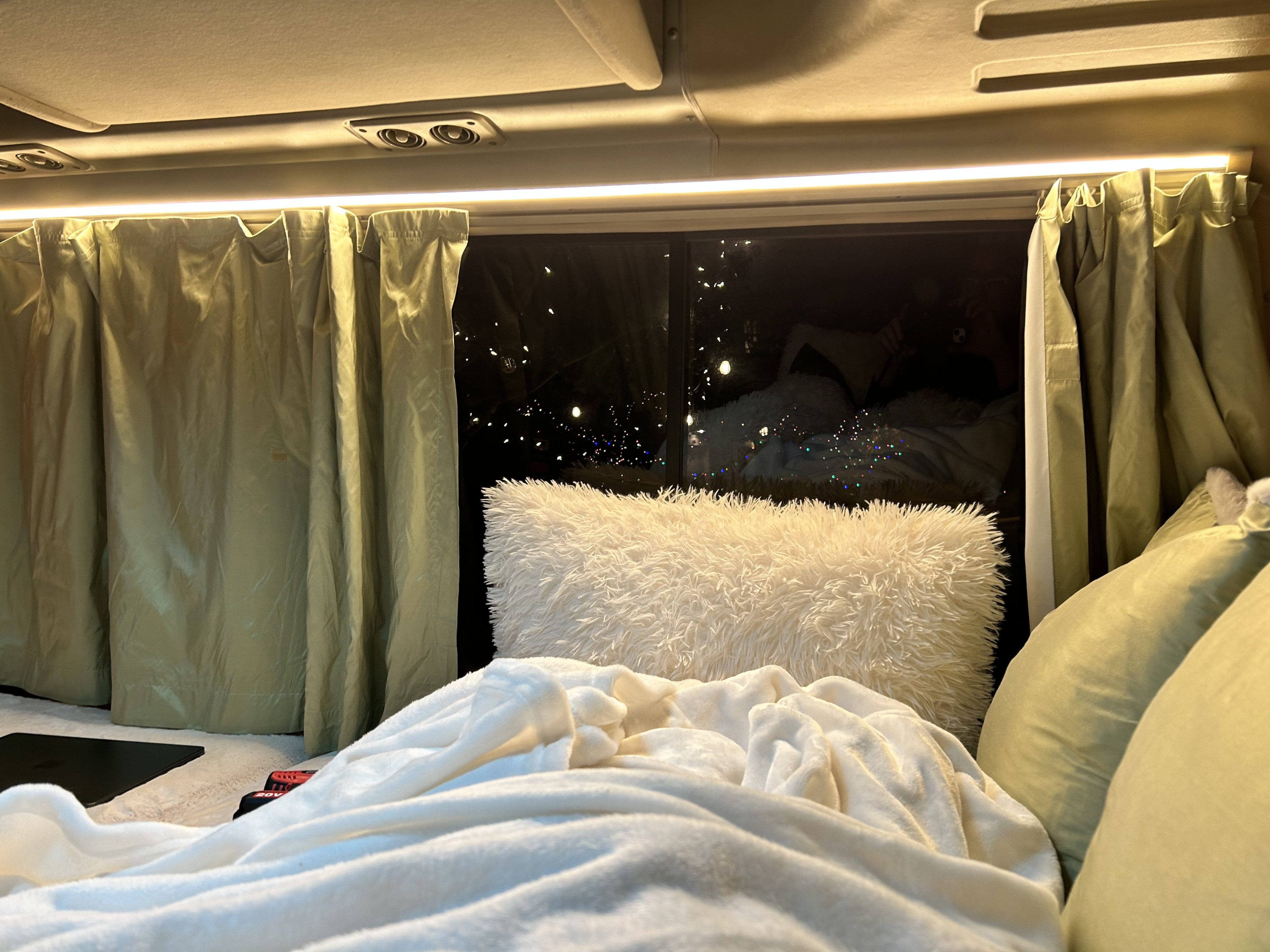 Cozy interior of an RV rental at night, showing a bed with fluffy pillows, green curtains, and blurred lights outside.. Toyota Coaster 1991