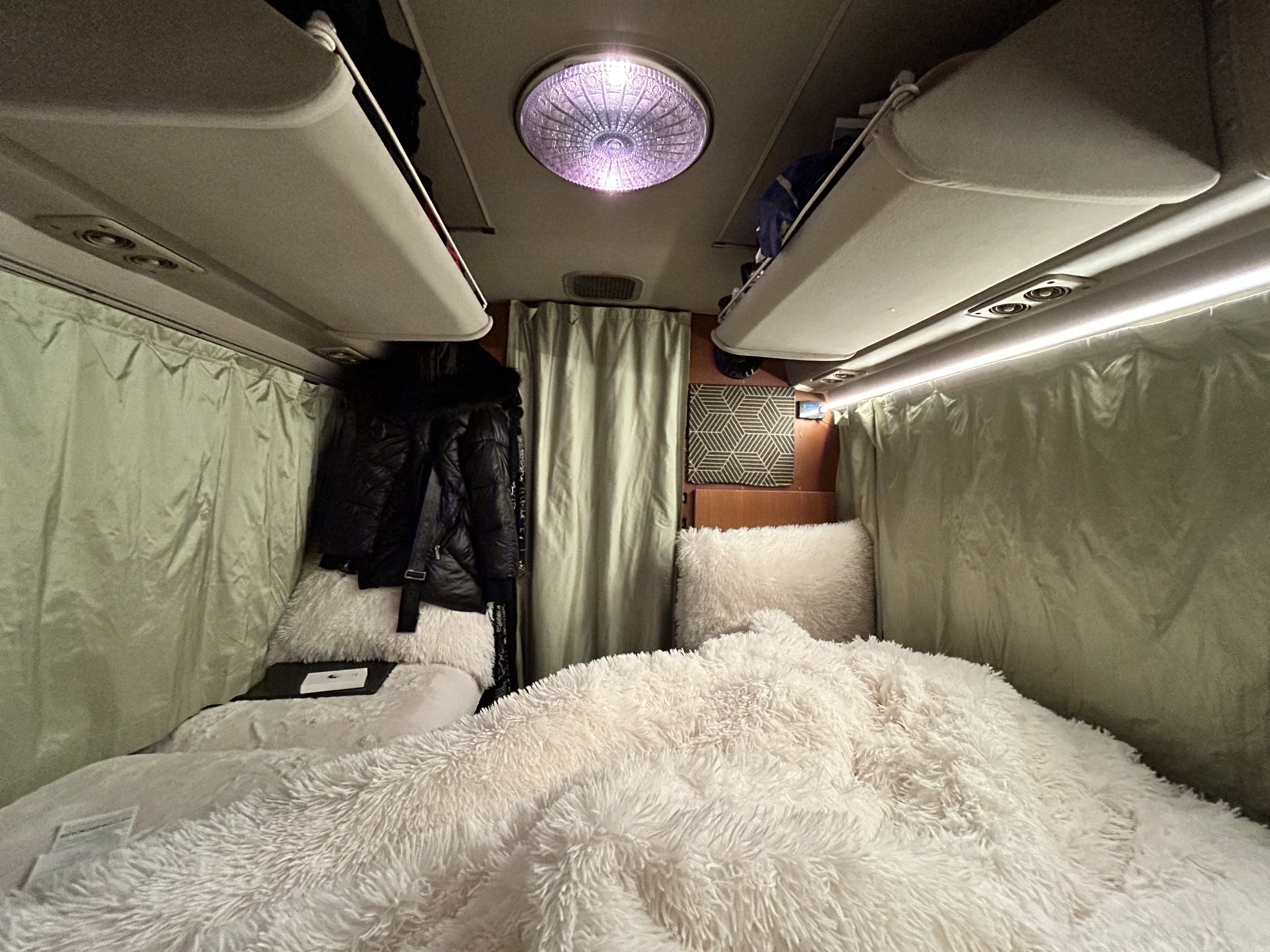 Cozy RV rental interior featuring a plush white bed, hanging clothes, curtains, and overhead compartments.. Toyota Coaster 1991