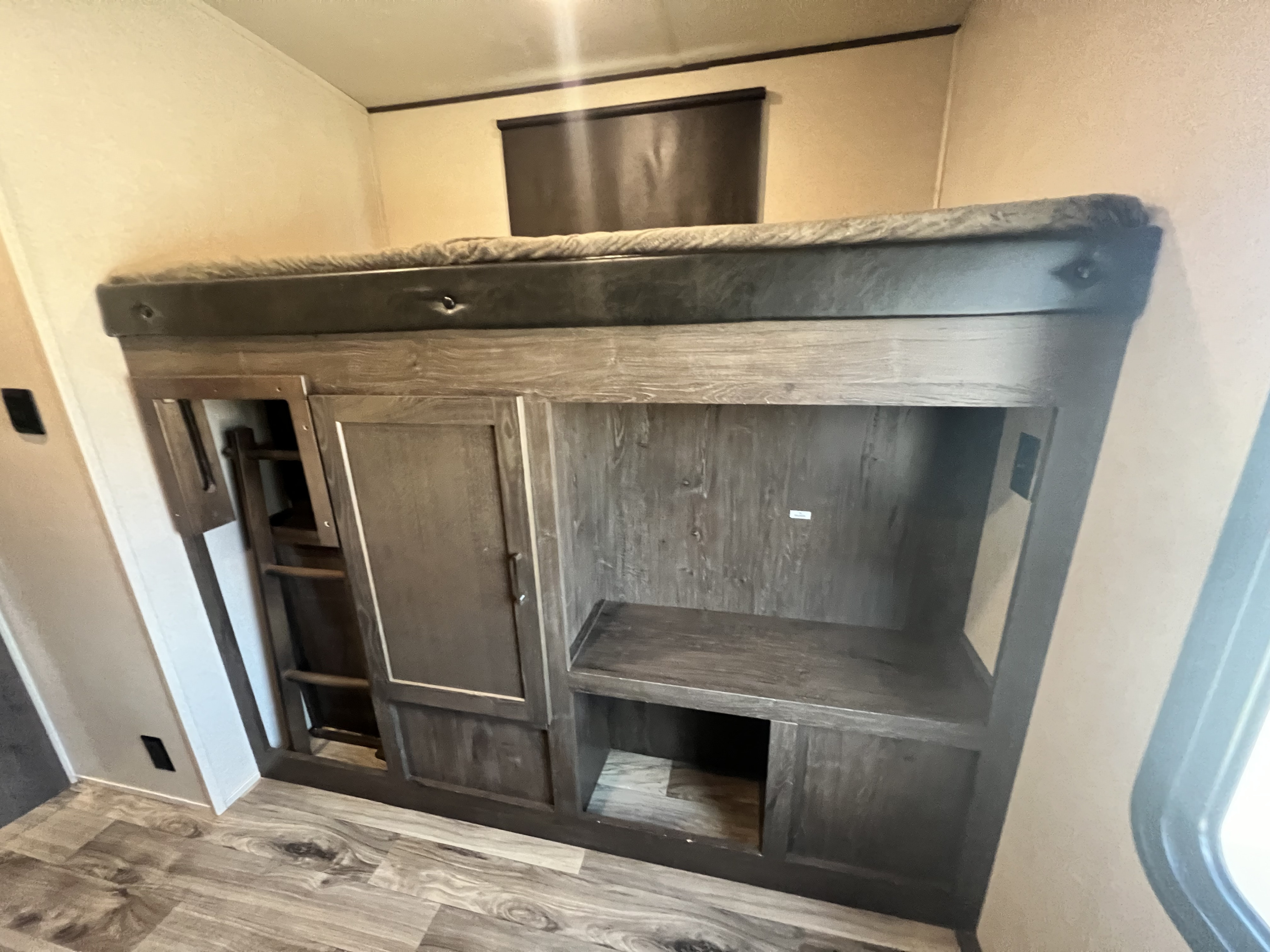 Inside an RV rental, a bunk bed with a mattress sits above integrated dark wood storage, shelves, and a stowed ladder.. Forest River Heritage Glen 2018