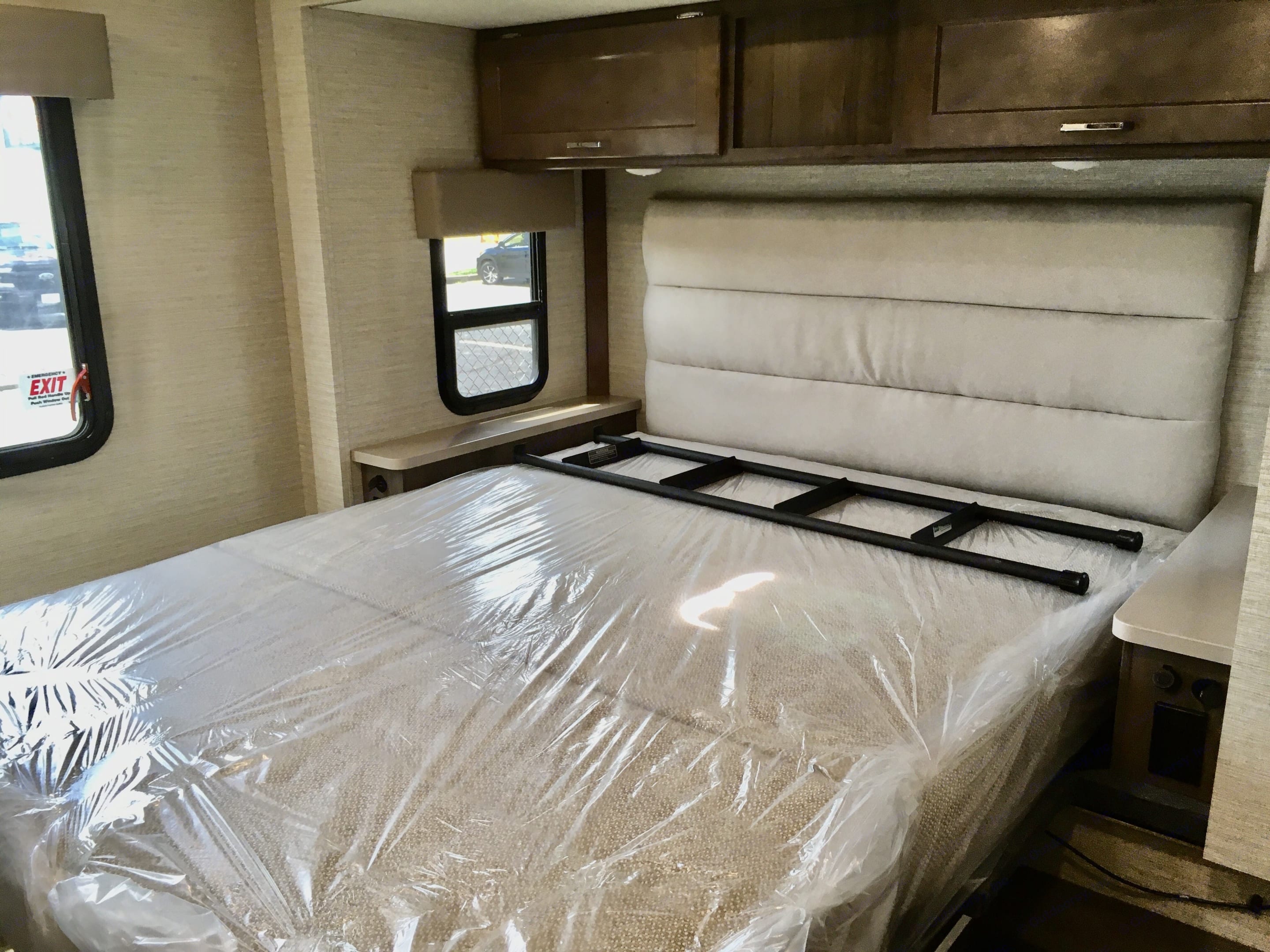 Inside an RV rental bedroom with a new, plastic-wrapped mattress, padded headboard, overhead cabinets, and side windows.. Winnebago Minnie Winnie 2024