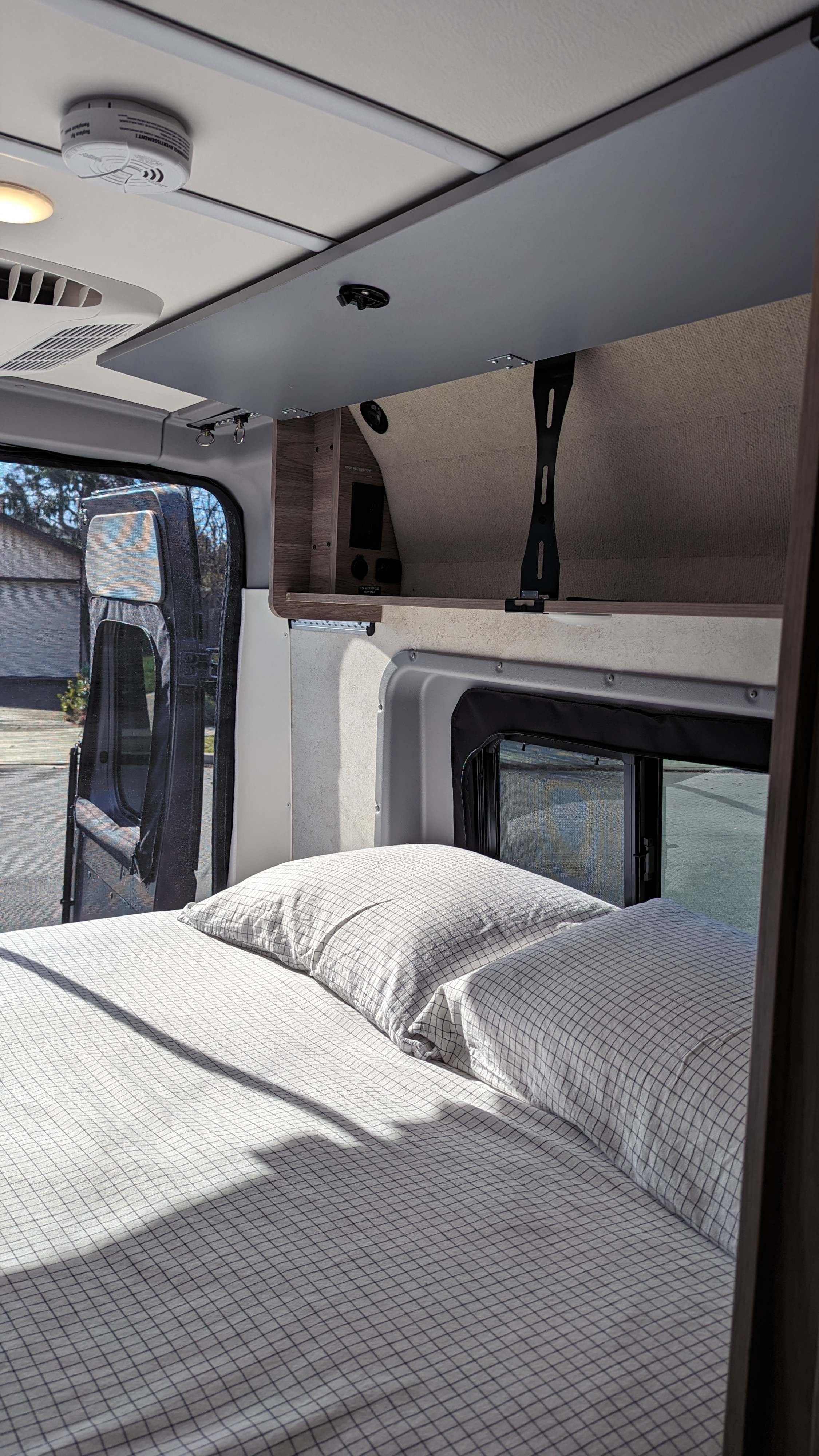 Inside an RV rental: a comfy bed with grid bedding, overhead storage, and sunlit windows revealing the outdoors.. Winnebago Solis 2023