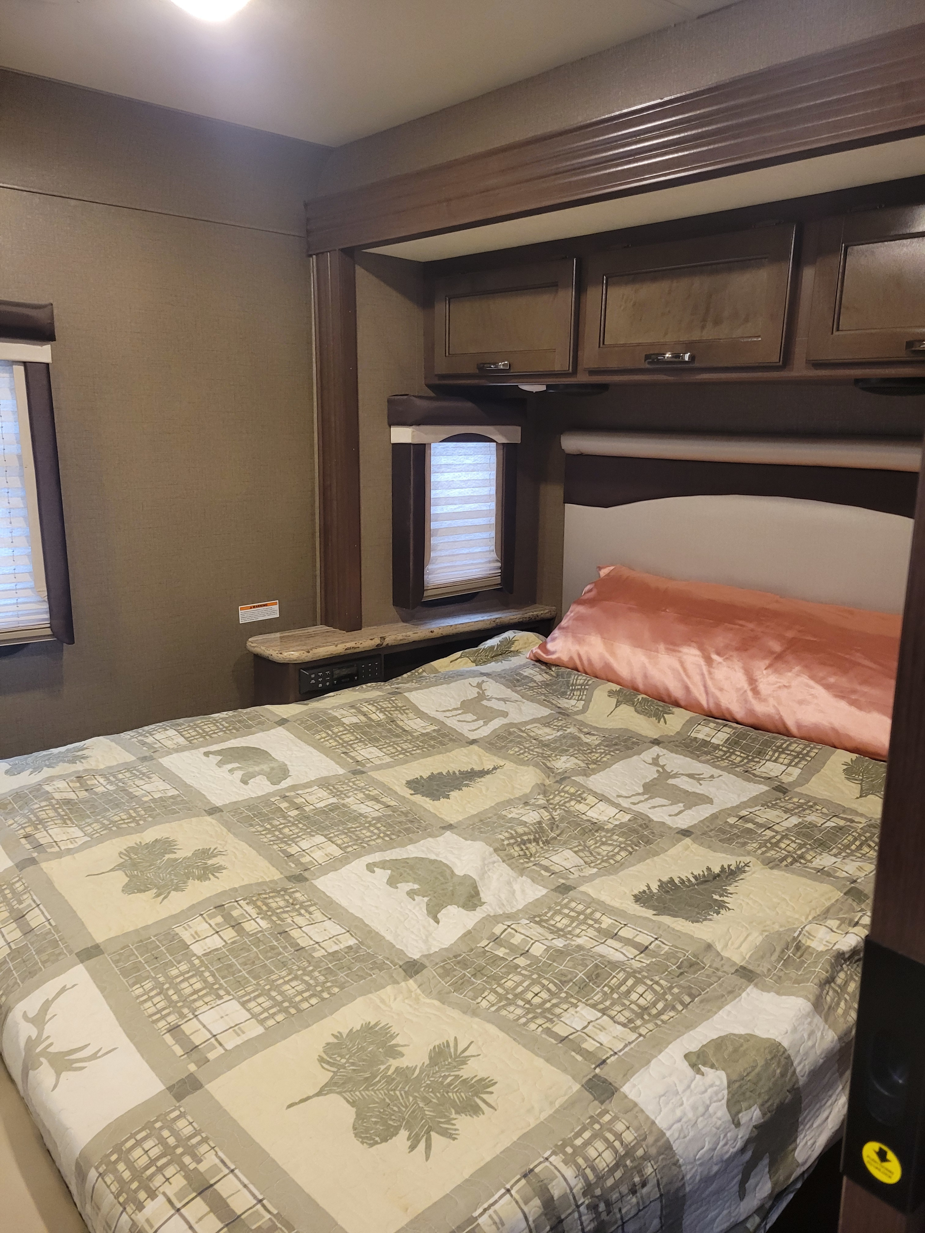 The bedroom of an RV rental, featuring a bed with a nature-themed quilt, compact cabinets, and integrated bedside controls.. Thor A.C.E. 2018