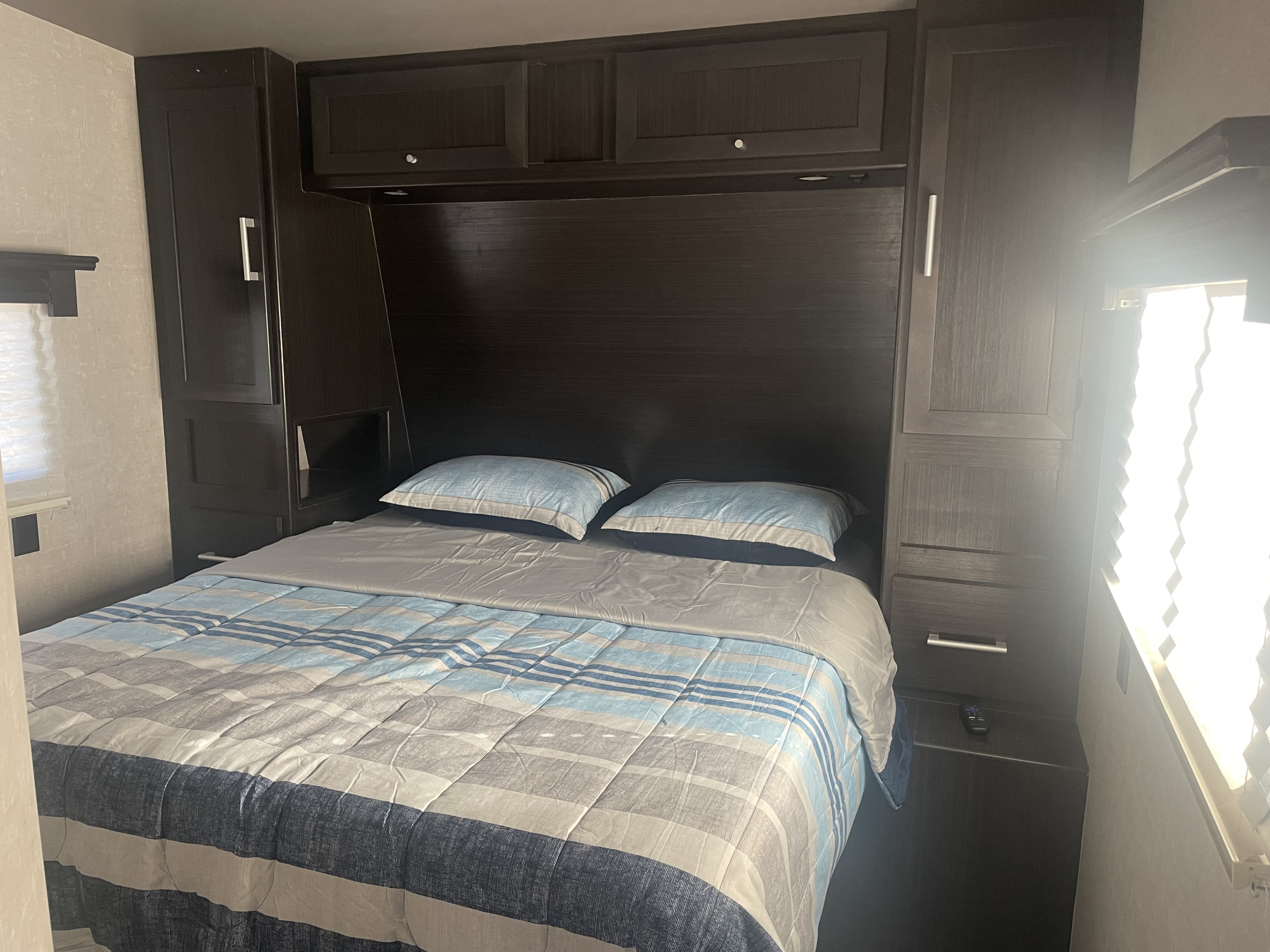 Cozy RV rental bedroom with dark wood cabinets and a striped comforter. Features a bed surrounded by practical storage, ideal for travel.. Eclipse Attitude Pro Lite Toy Hauler 2019