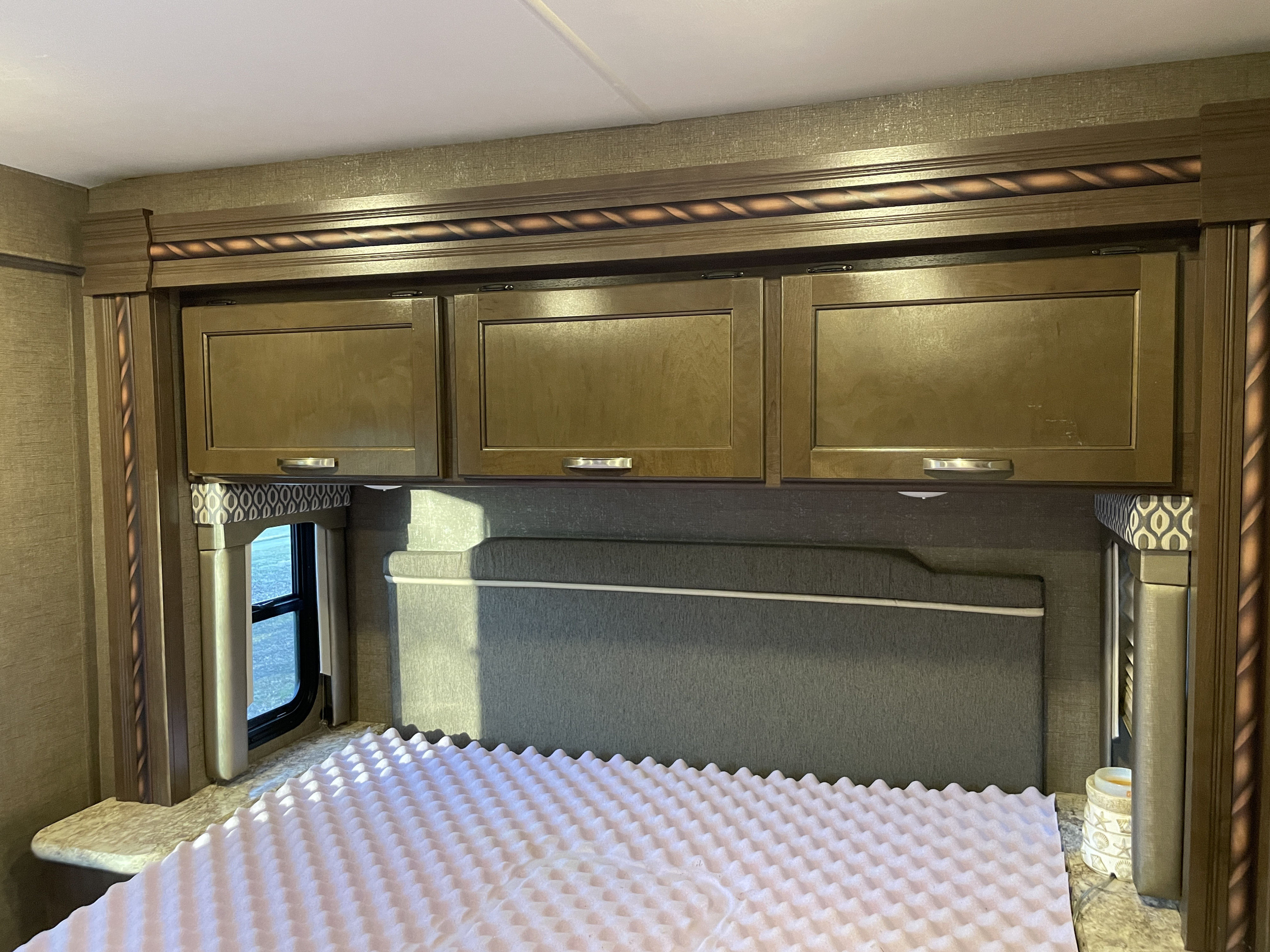 Interior of an RV rental bedroom showing an unmade bed with a foam topper, a dark headboard, and wood overhead cabinets.. Thor Freedom Elite 2017