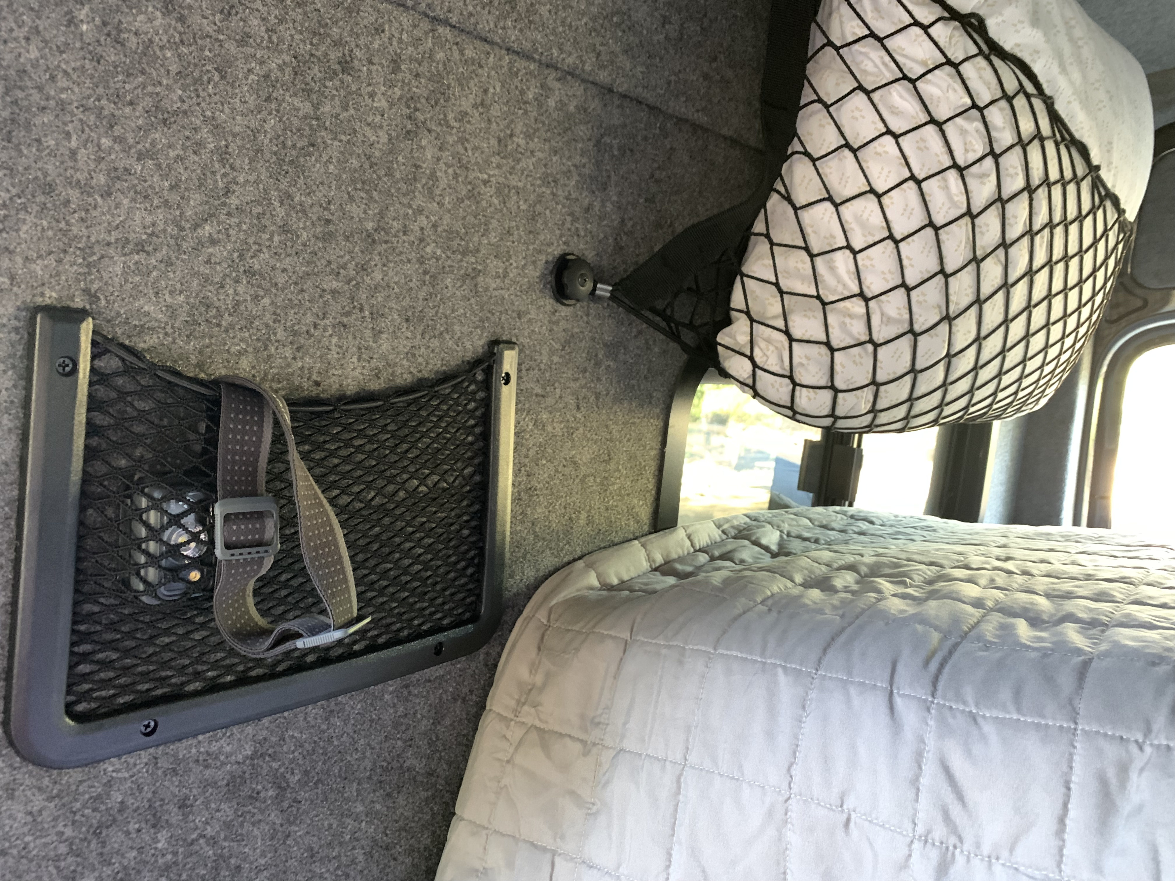 Practical RV rental interior: soft bed, wall-mounted mesh storage with headlamp, and an overhead net for pillows.. Mercedes-Benz Sprinter 2016