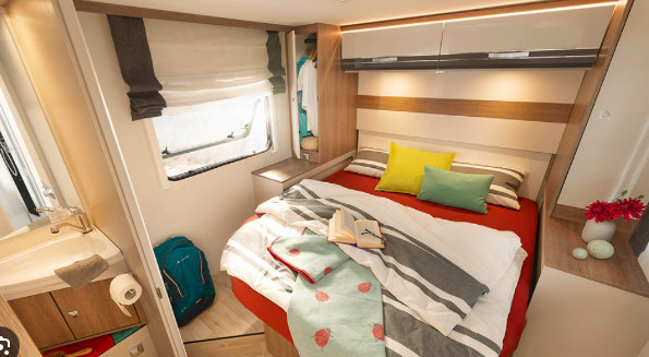 This RV rental interior shows a compact bedroom with a comfy bed, storage, window, and a small bathroom area.. Dethleffs Trend 90 T 7057DBL 2024