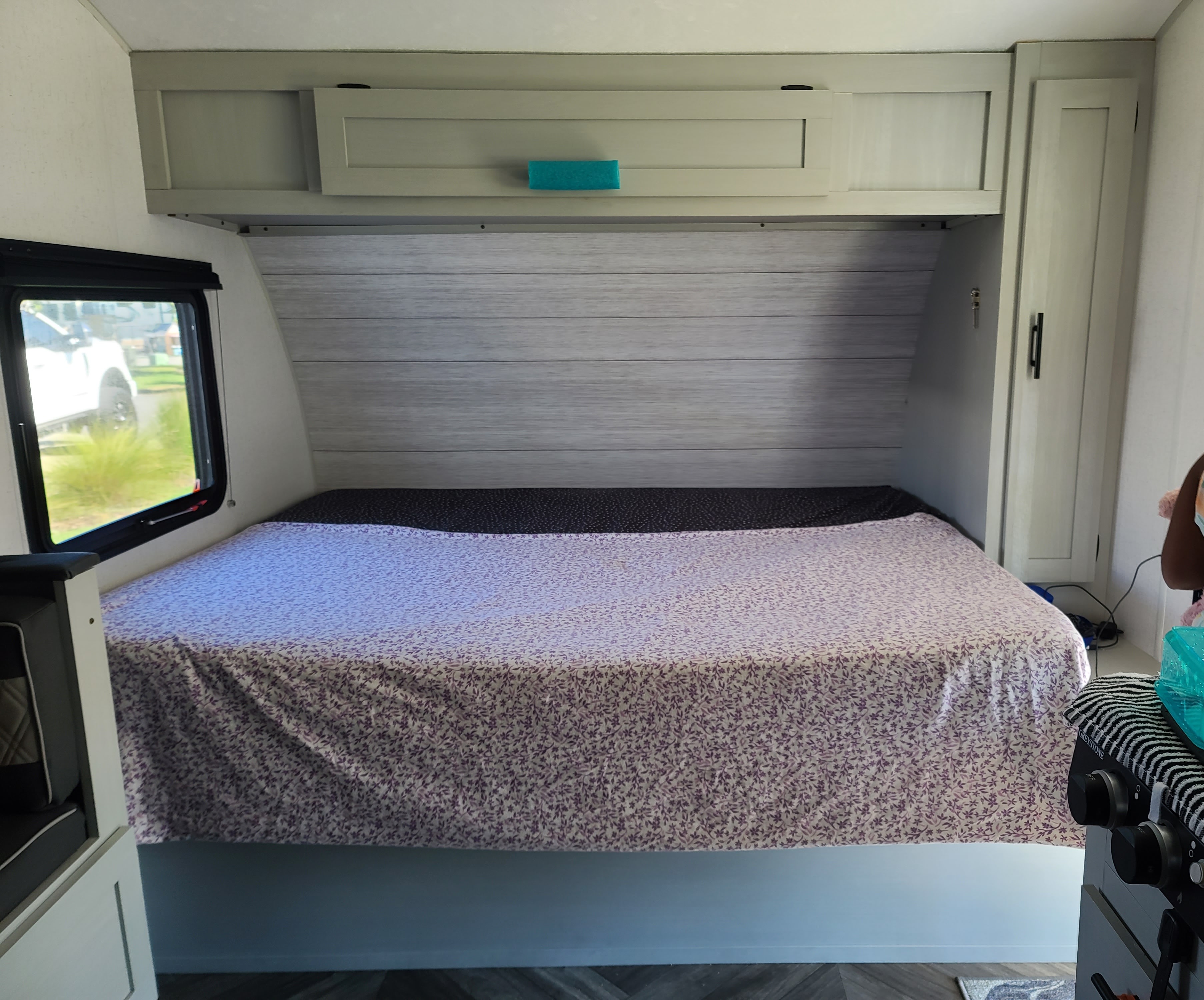 An RV rental bedroom shows a bed with purple patterned bedding, overhead storage cabinets, and a window for natural light.. Forest River Salem FSX 2022