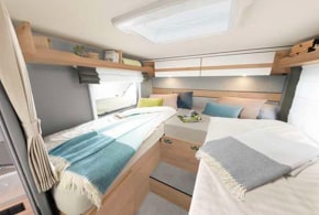 Inside an RV rental: a bright, comfortable bedroom with two beds, overhead storage, and natural wood accents, perfect for restful travel.. Dethleffs Just T 7052EBL 2024