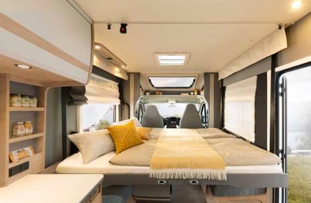 An inviting RV rental interior featuring a large bed with yellow accents, a built-in shelf, and the driver's cabin visible in the background.. Dethleffs Just T 7052EBL 2024