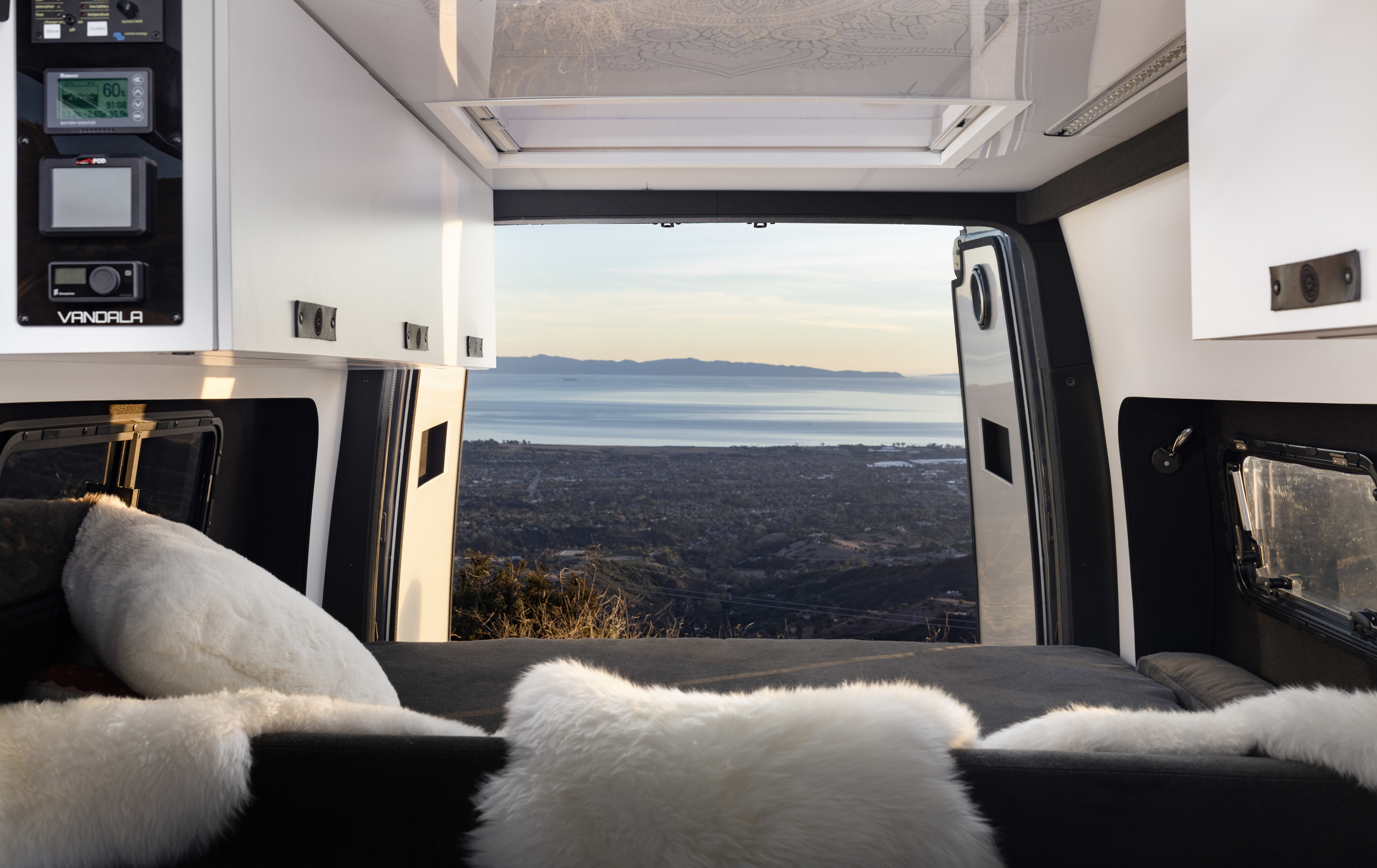 Cozy camper van RV rental interior with fluffy bedding and an expansive view of mountains, town, and ocean from the open rear doors.. Mercedes-Benz Sprinter 144 High Roof 2022