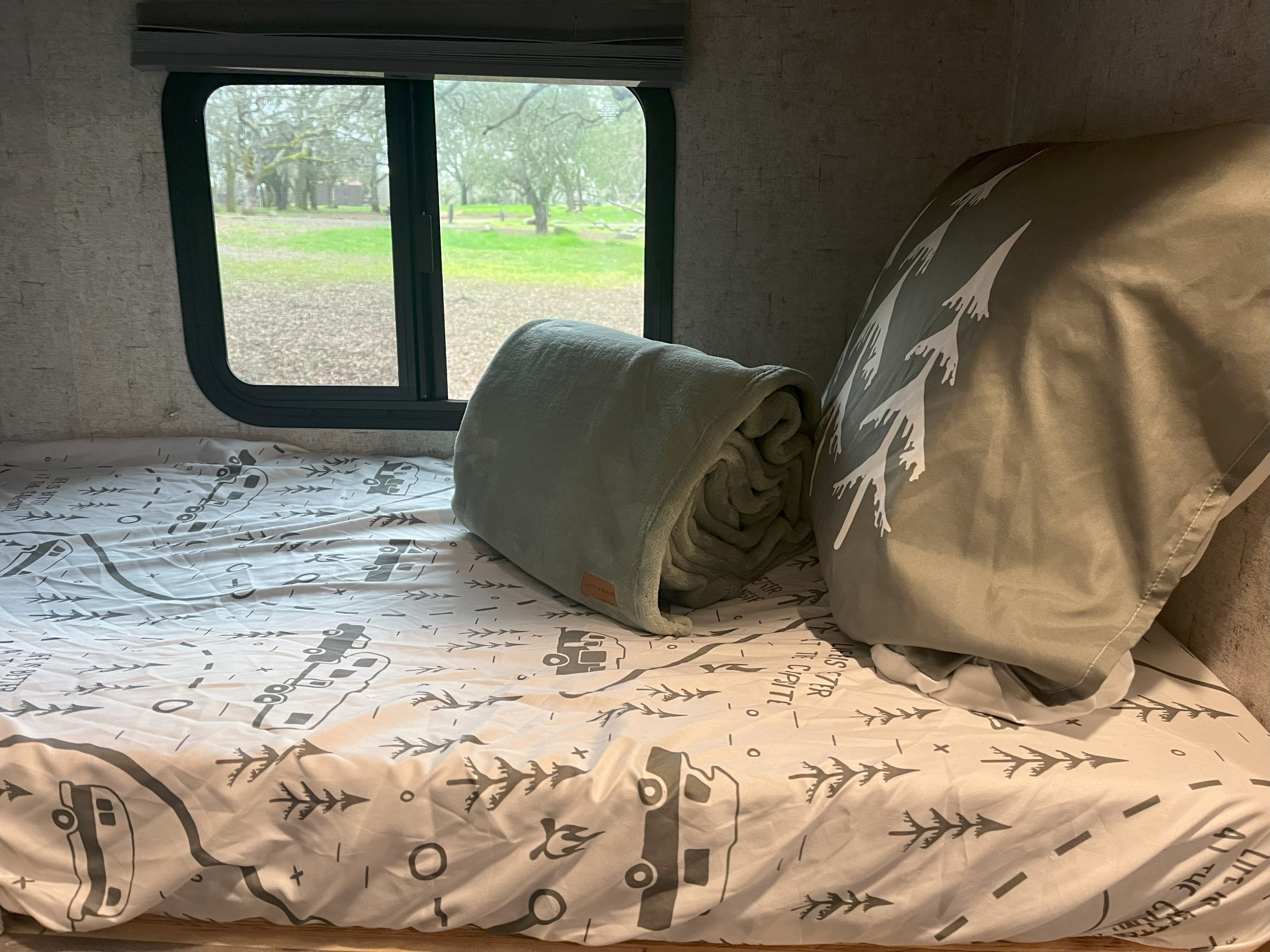 Cozy RV rental bunk with patterned bedding, a blanket, and pillow, overlooking a green campsite through the window.. Coachmen Apex Nano 2019