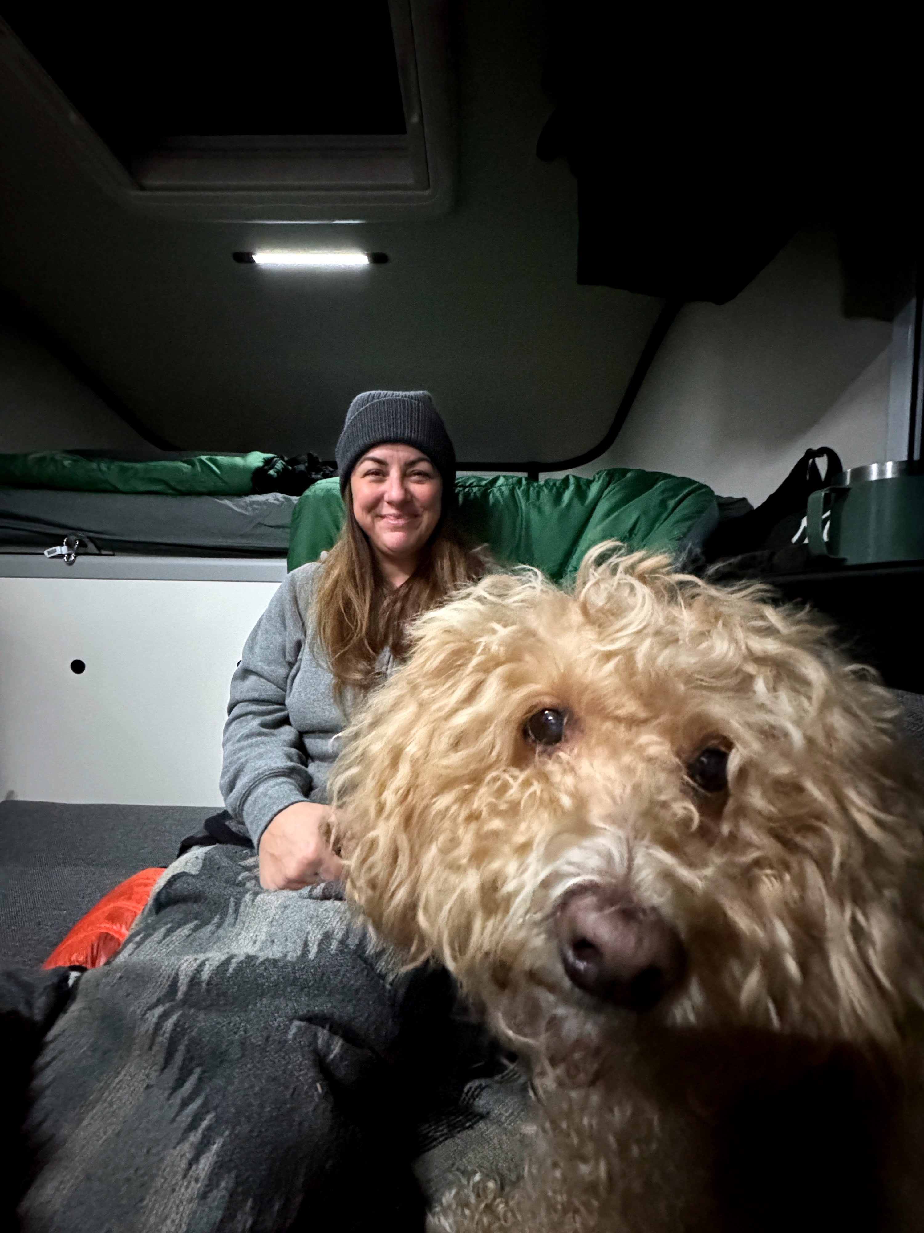 A smiling woman and fluffy dog are cozy inside an RV rental. Green bedding and a shelf visible in the camper interior.. Dodge Dodge 1 Ton Pu 2018