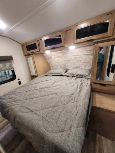 This RV rental features a comfortable bedroom with overhead storage, a cozy bed, and modern wood accents.. Forest River 26 DBH 2024