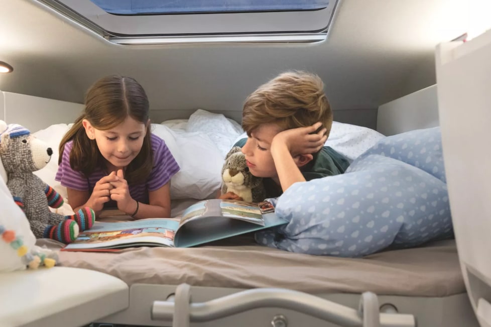 Two kids with stuffed animals read in a cozy RV rental bunk. Perfect for family adventures on the road!. Volkswagen Grand California 600 boîte automatique 2024
