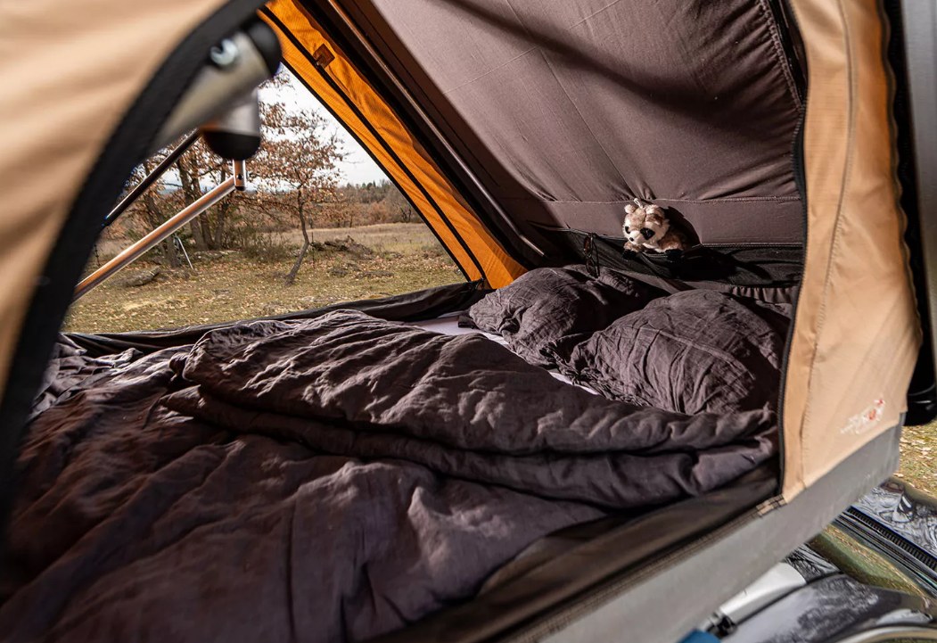 Cozy interior of a rooftop tent with dark bedding and a stuffed animal, offering comfort for an RV rental camping adventure in nature.. NaitUp Duo 2021