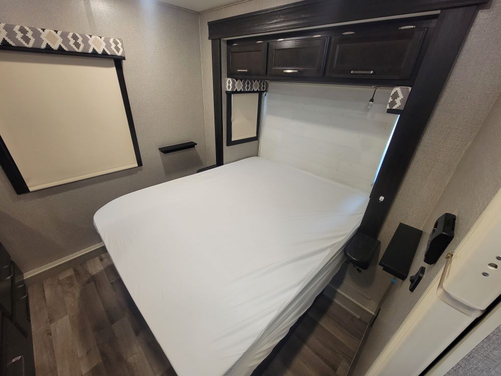 Compact RV rental bedroom with white bed, window shade, overhead cabinets, and wood-look floor.. Jayco Alante 31V 2022