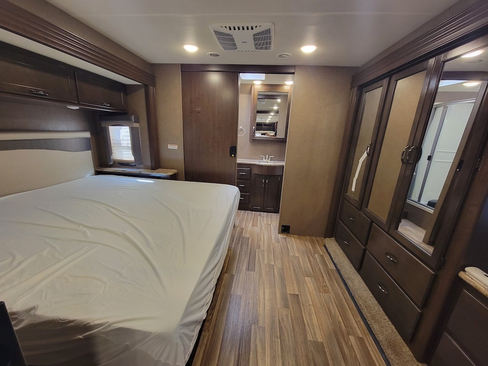 Inside an RV rental: a modern bedroom with a comfy bed, wood floors, dark wood cabinets, and an attached bath.. Thor A.C.E. 2019