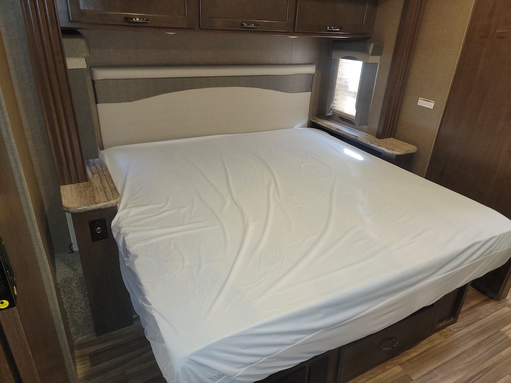 RV rental bedroom showcasing a bed with a white cover, headboard, overhead cabinets, and compact side tables.. Thor A.C.E. 2019